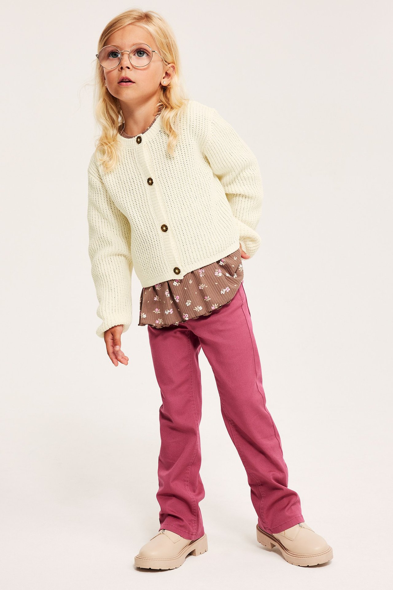 Frontvisning: Barn i hvit strikket cardigan med brune knapper, blomstret topp, rosa bukser og beige sko.