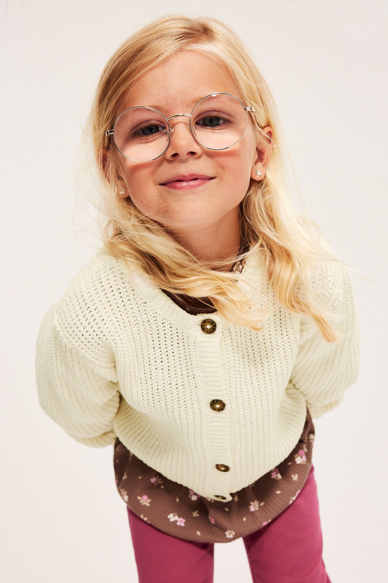 Frontvisning: Barn i hvit strikket cardigan med brune knapper, brun blomstret topp, rosa leggings og runde briller.