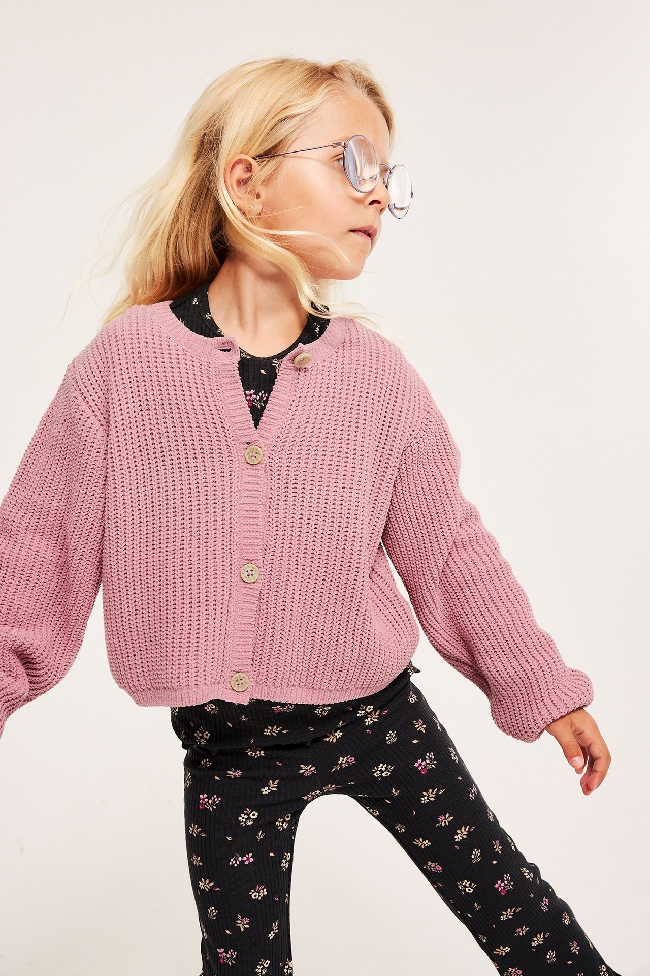 Forfra: Barn i rosa strikket cardigan med knapper over svart blomstret topp og leggings, med briller.