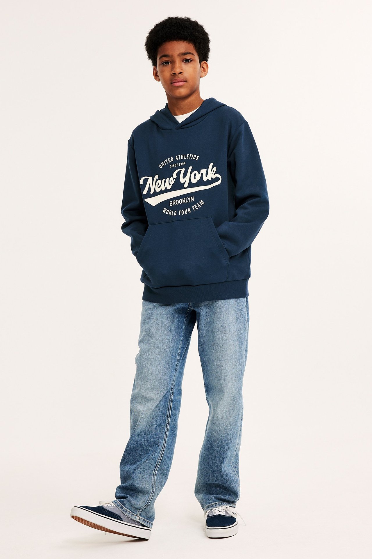 Edestä kuvattu lapsi tummansinisessä New York -hupparissa, vaaleansiniset farkut ja tummansiniset tennarit.