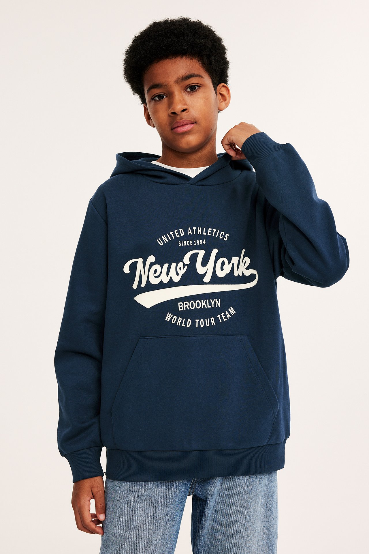 Frontvisning av et barn i en mørkeblå hettegenser med hvitt 'New York'-trykk og lyseblå jeans.