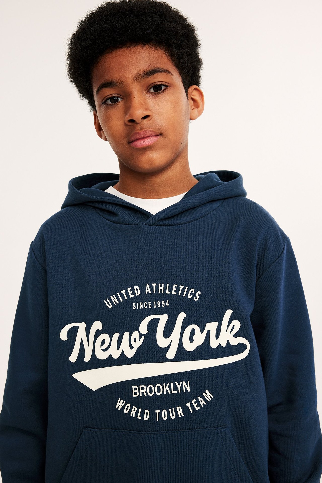 Edestä kuvattu lapsi tummansinisessä hupparissa, jossa valkoinen "New York" -painatus ja kengurutasku.