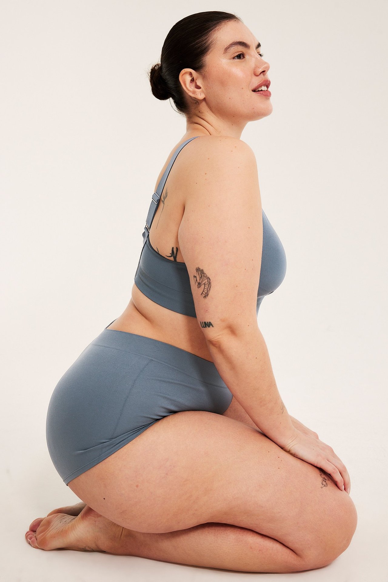 Sivukuva polvistuvasta naisesta tummansinisissä saumattomissa bokserialushousuissa ja yhteensopivissa rintaliiveissä.