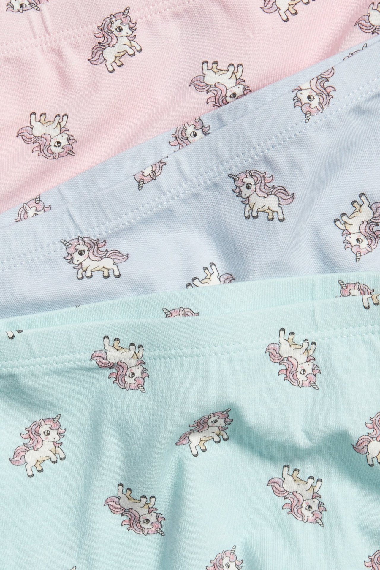 Zbliżenie na trzy pary bokserek dziecięcych w kolorze różowym, jasnoniebieskim i miętowym z wzorem jednorożców.