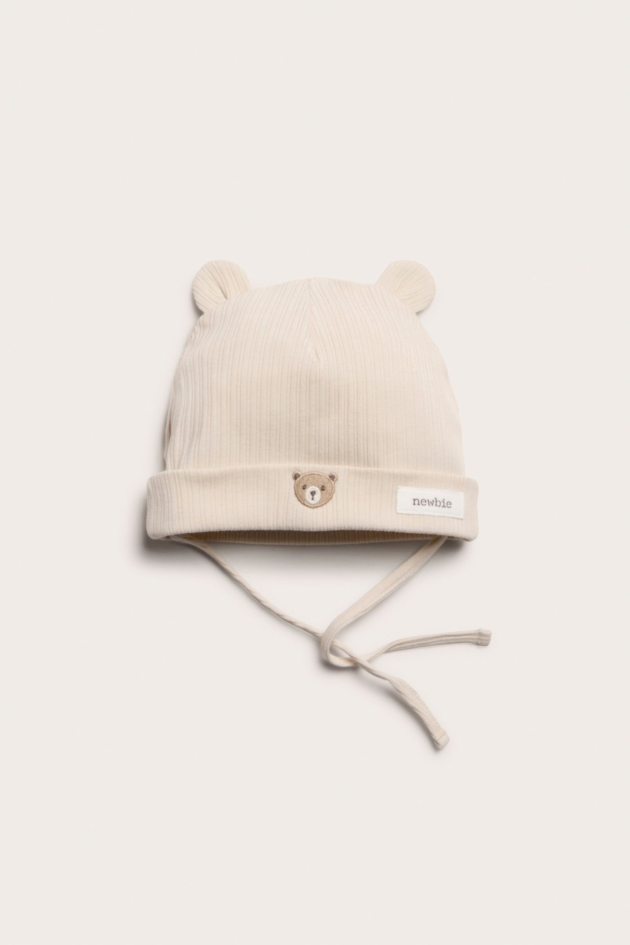 Widok z przodu jasnobeżowej prążkowanej czapeczki niemowlęcej z uszami misia, haftowanym misiem i wiązaniami.