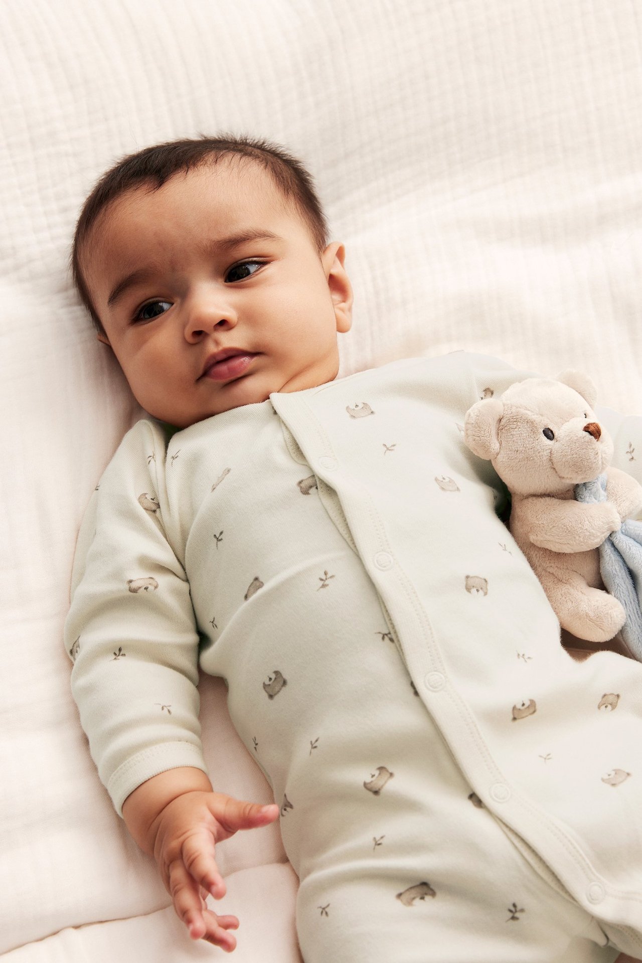 Närbild på baby i ljusbeige bomullspyjamas med nallemönster, tryckknappar, bredvid en nallebjörn.