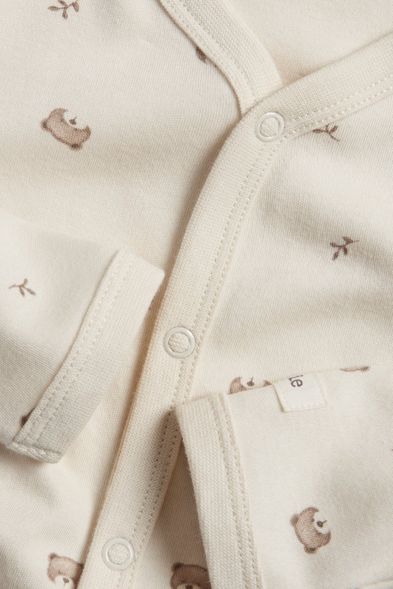 Närbild av en ljusbeige pyjamas i bomull för baby med brunt nallemönster och tryckknappar.