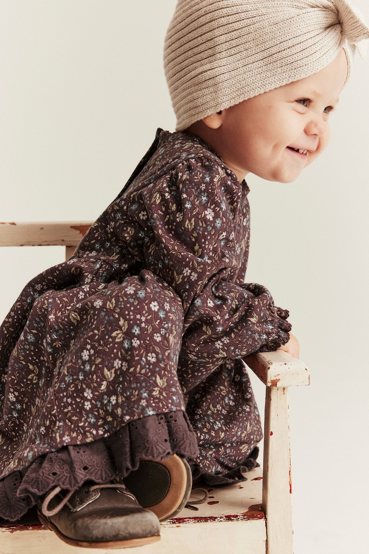 Sidovy av baby i mörklila blommig jerseyklänning med spetsfåll, beige stickad turban och bruna skor.