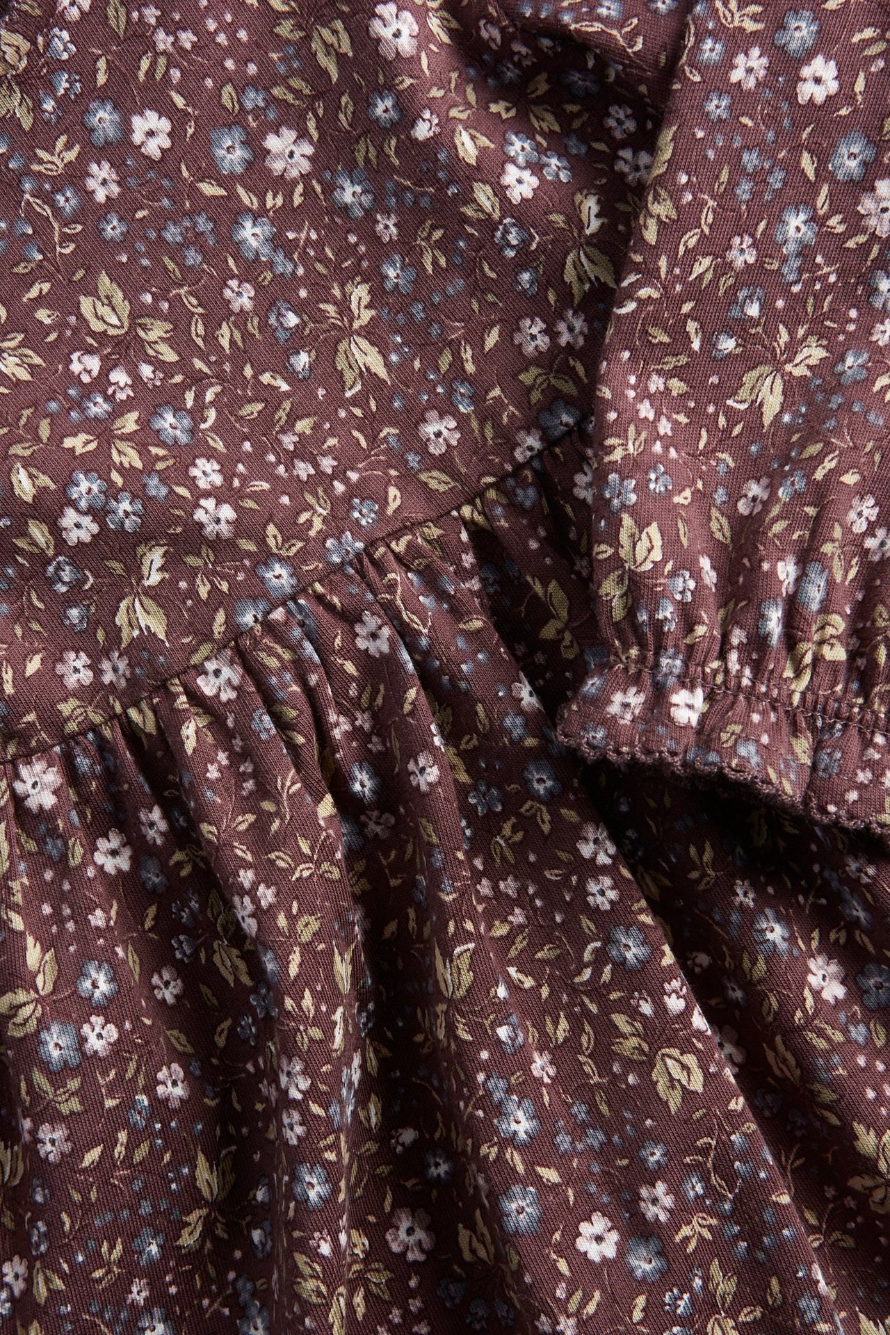 Zbliżenie liliowej sukienki dżersejowej w kwiaty z małymi białymi i niebieskimi kwiatami, falbankami i elastycznym mankietem.