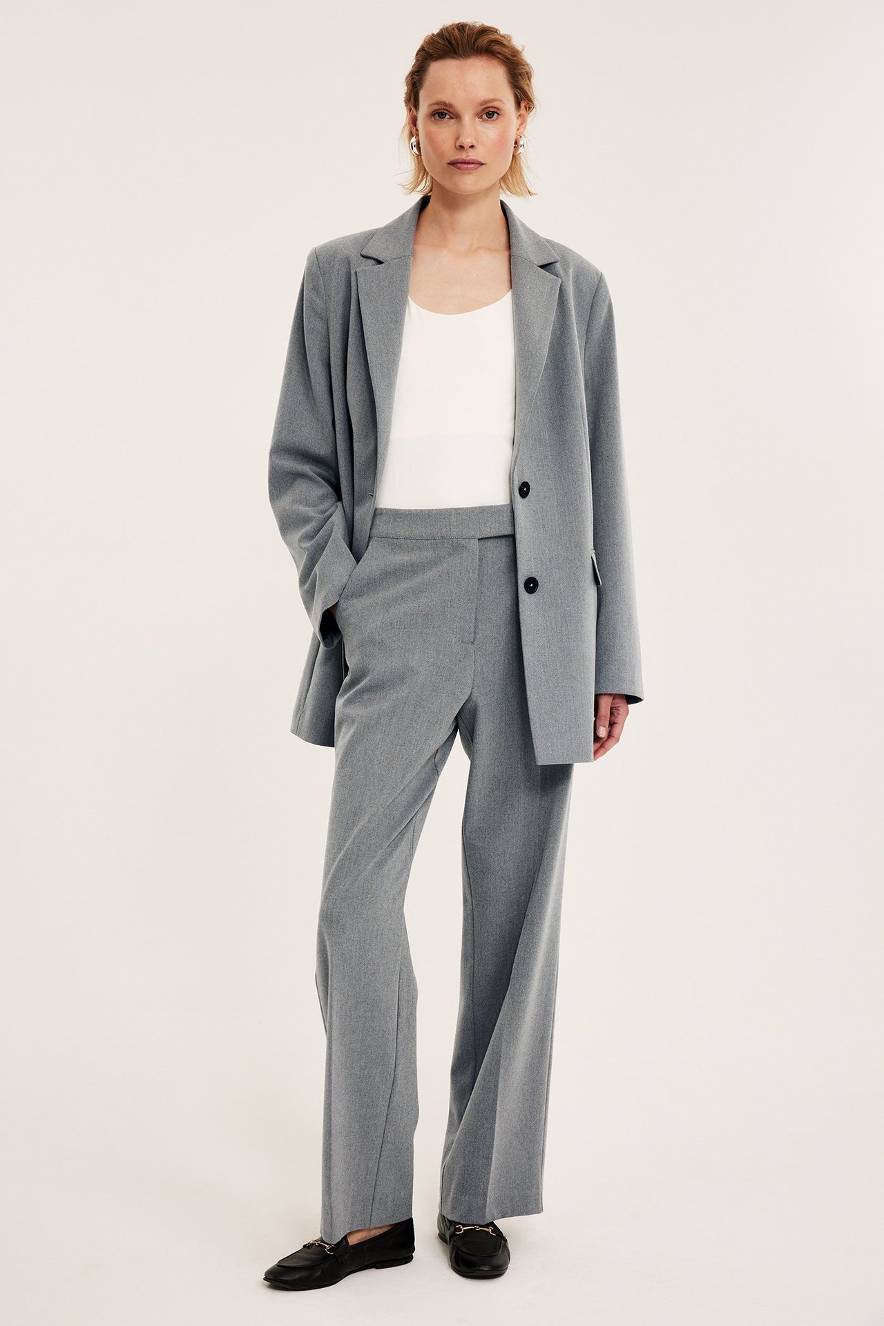 Frontvisning: Kvinne i lys grå melert dressbukse, matchende blazer, hvit topp og svarte loafers.
