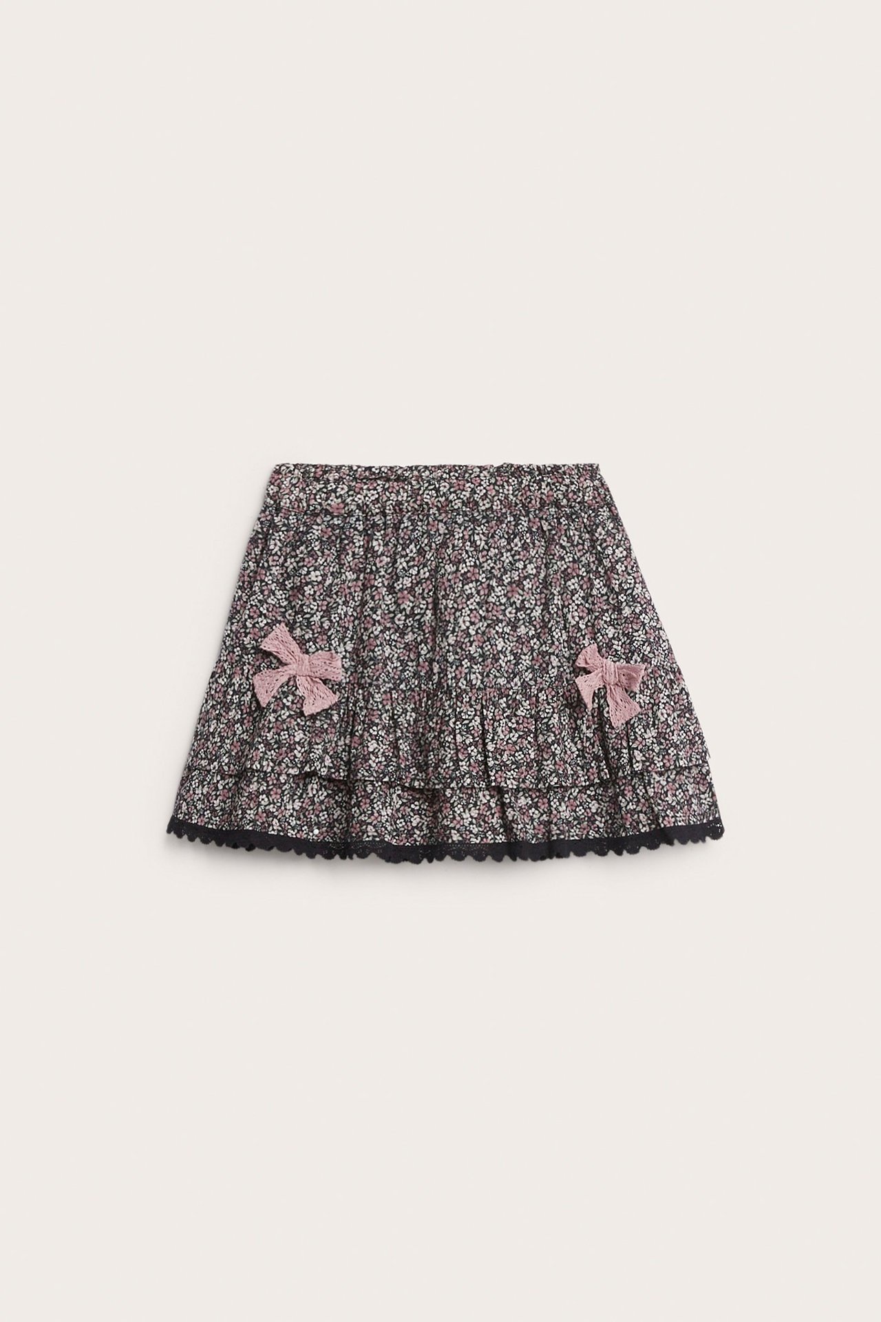 Frontbilde av et sort blomstret bomullsskjørt for barn med volanger, rosa sløyfer og sort blondekant.