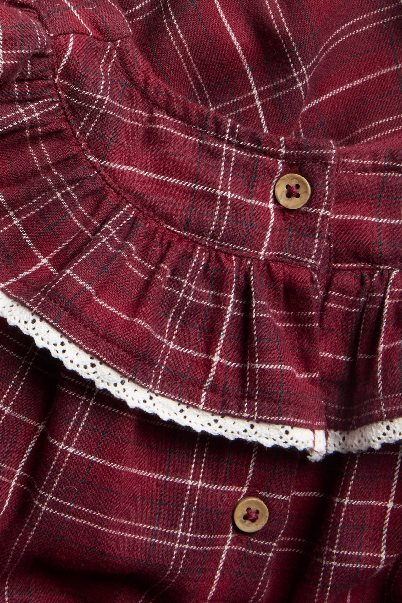 Zbliżenie czerwonej, kratkowanej bawełnianej sukienki z zaokrągloną falbanką, białą koronką i drewnianymi guzikami.