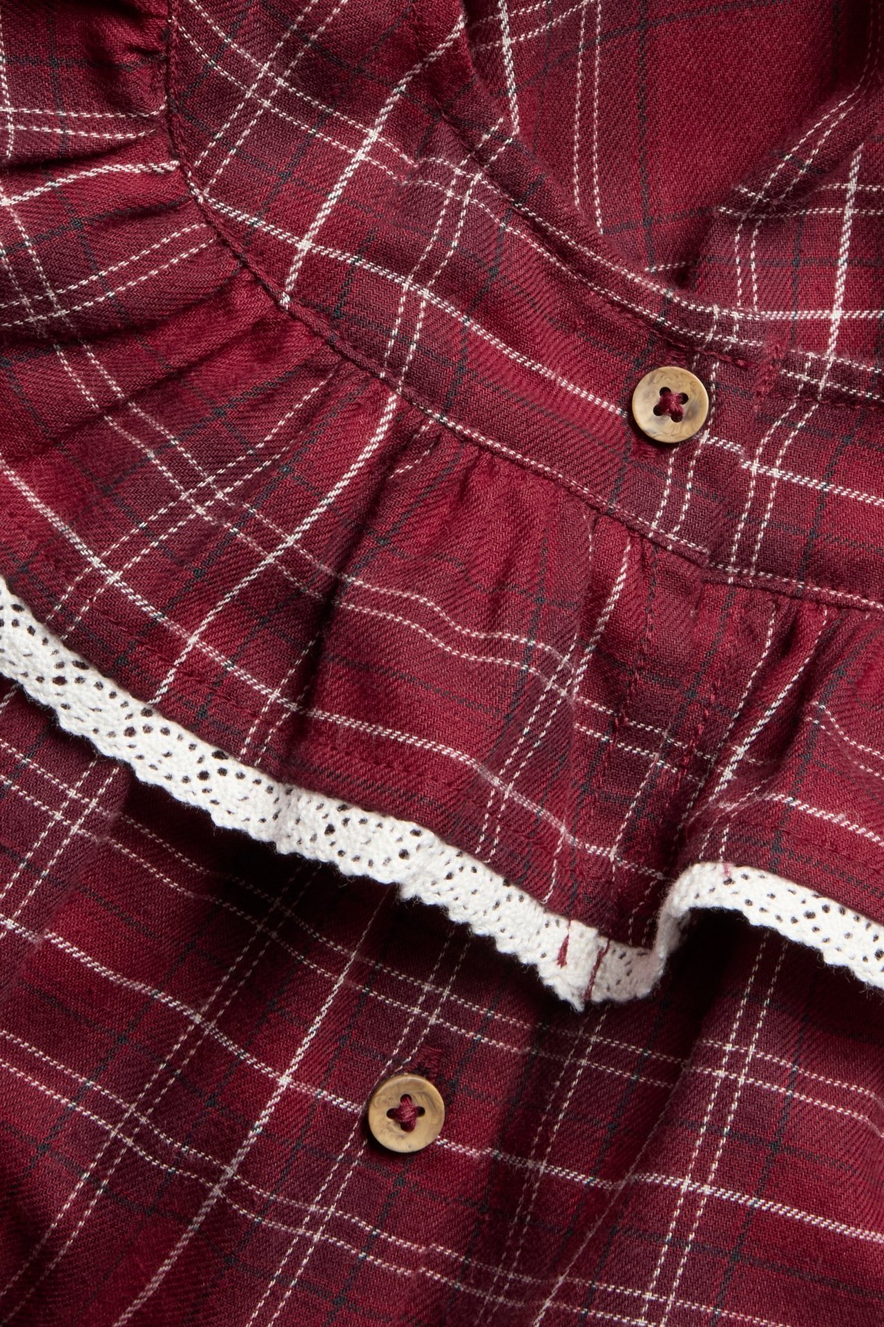 Zbliżenie na czerwoną sukienkę w kratę z zaokrągloną falbanką, białą koronką i dwoma drewnianymi guzikami.