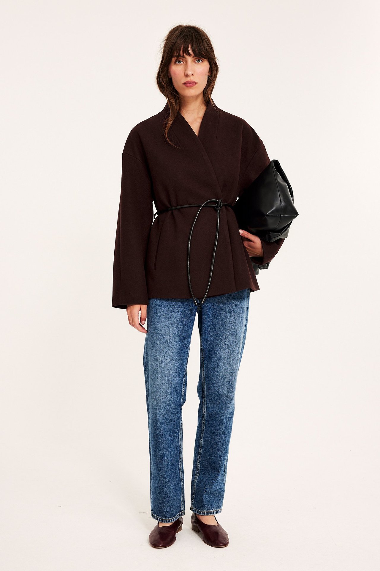 Forfra: Kvinne i vinrød kimonojakke, svart belte, blå jeans, mørkerøde ballerinasko, holder svart håndveske.