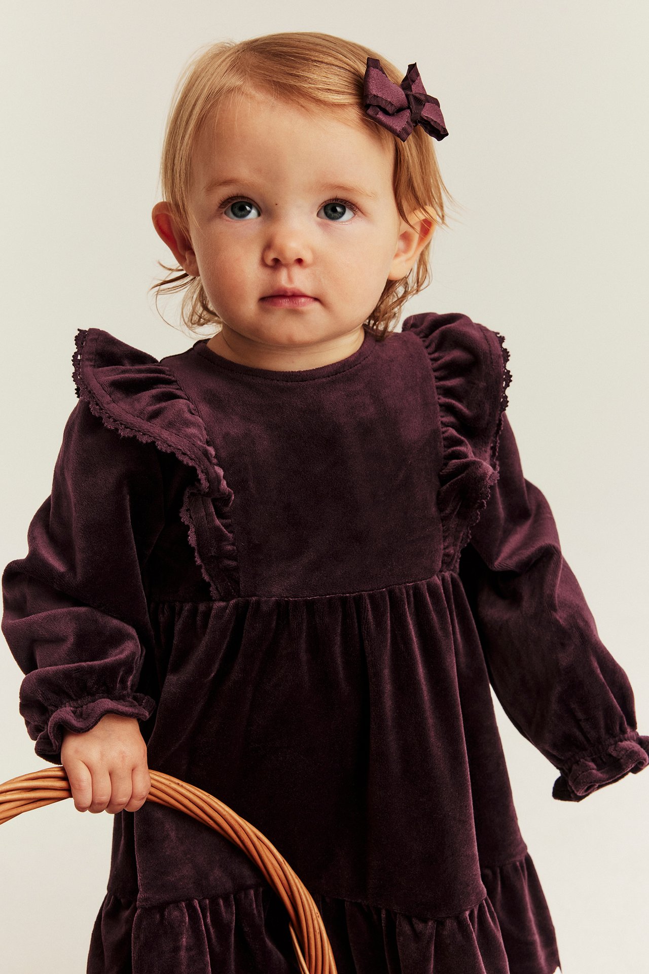 Framsida av en babyflicka i mörklila sammetsklänning med volanger och matchande rosett, håller en flätad korg.
