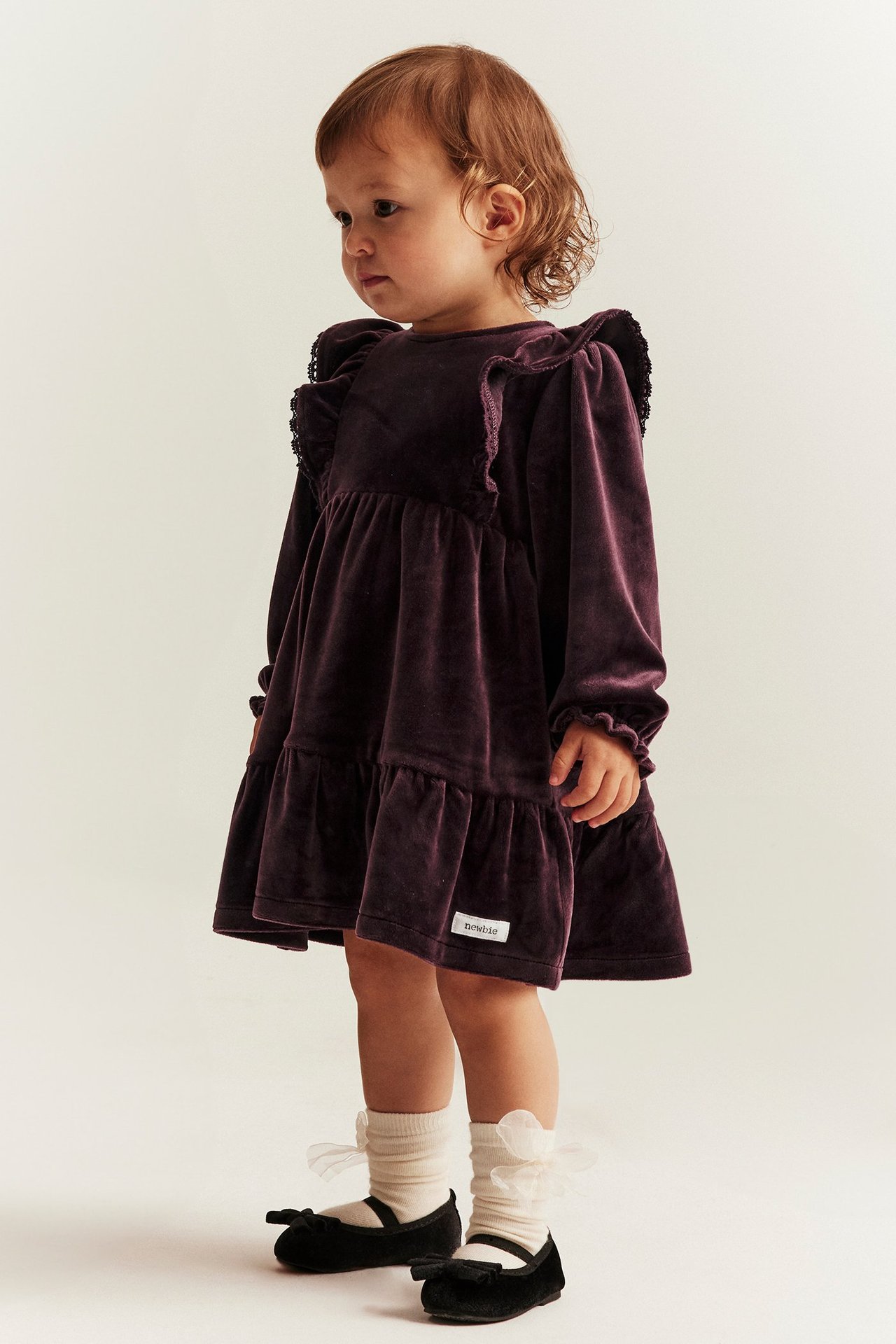 Lila velourklänning i recycled polyester med volanger, spetskant på axlar och bakknäppning.