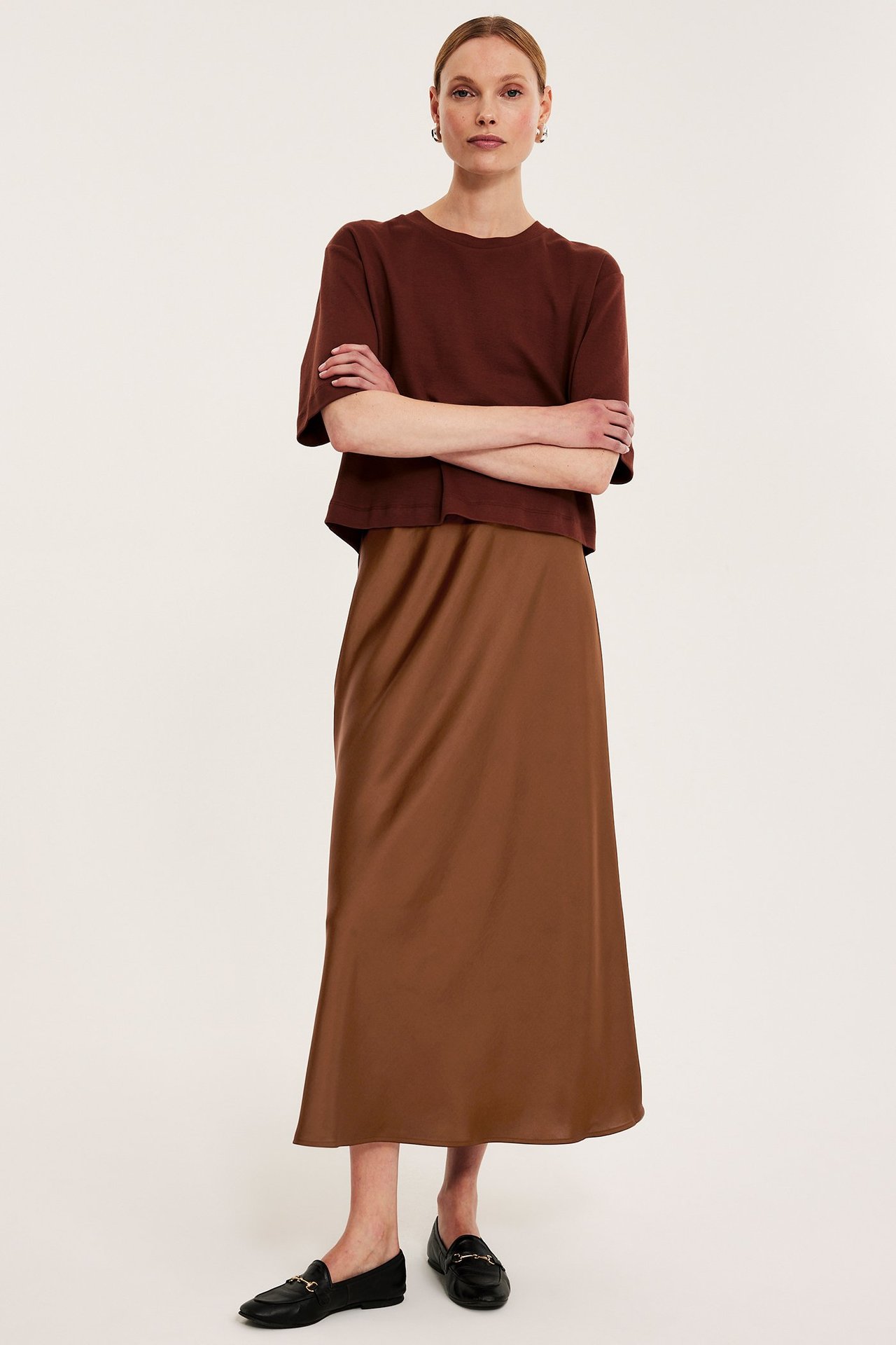 Edestä kuvattu nainen ruskeassa levenevässä satiinihameessa, yhdistettynä ruskeaan t-paitaan ja mustiin loafer-kenkiin.