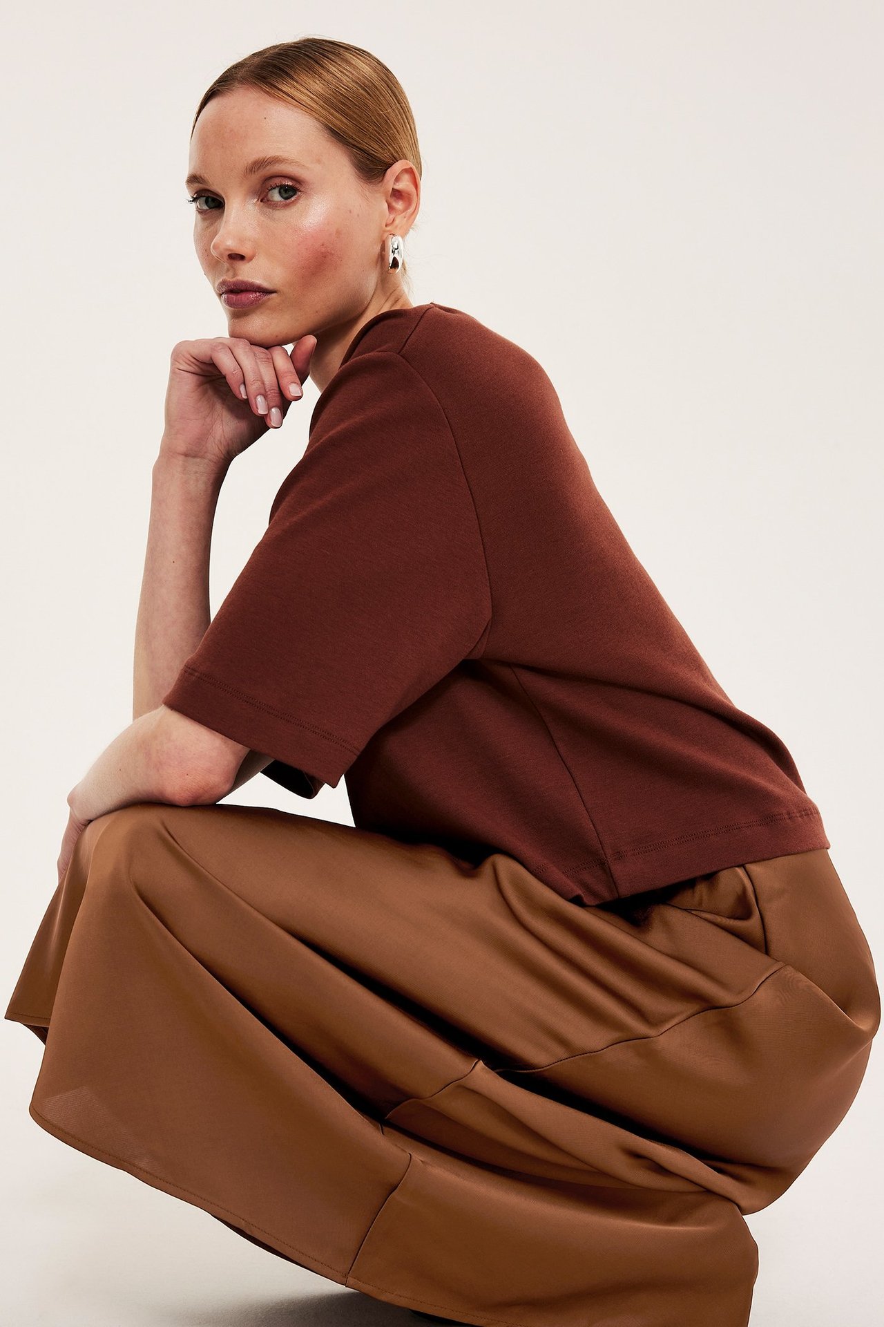 Sivukuva naisesta ruskeassa satiinihameessa, ruskeassa lyhythihaisessa topissa ja hopeisissa korvarenkaissa.