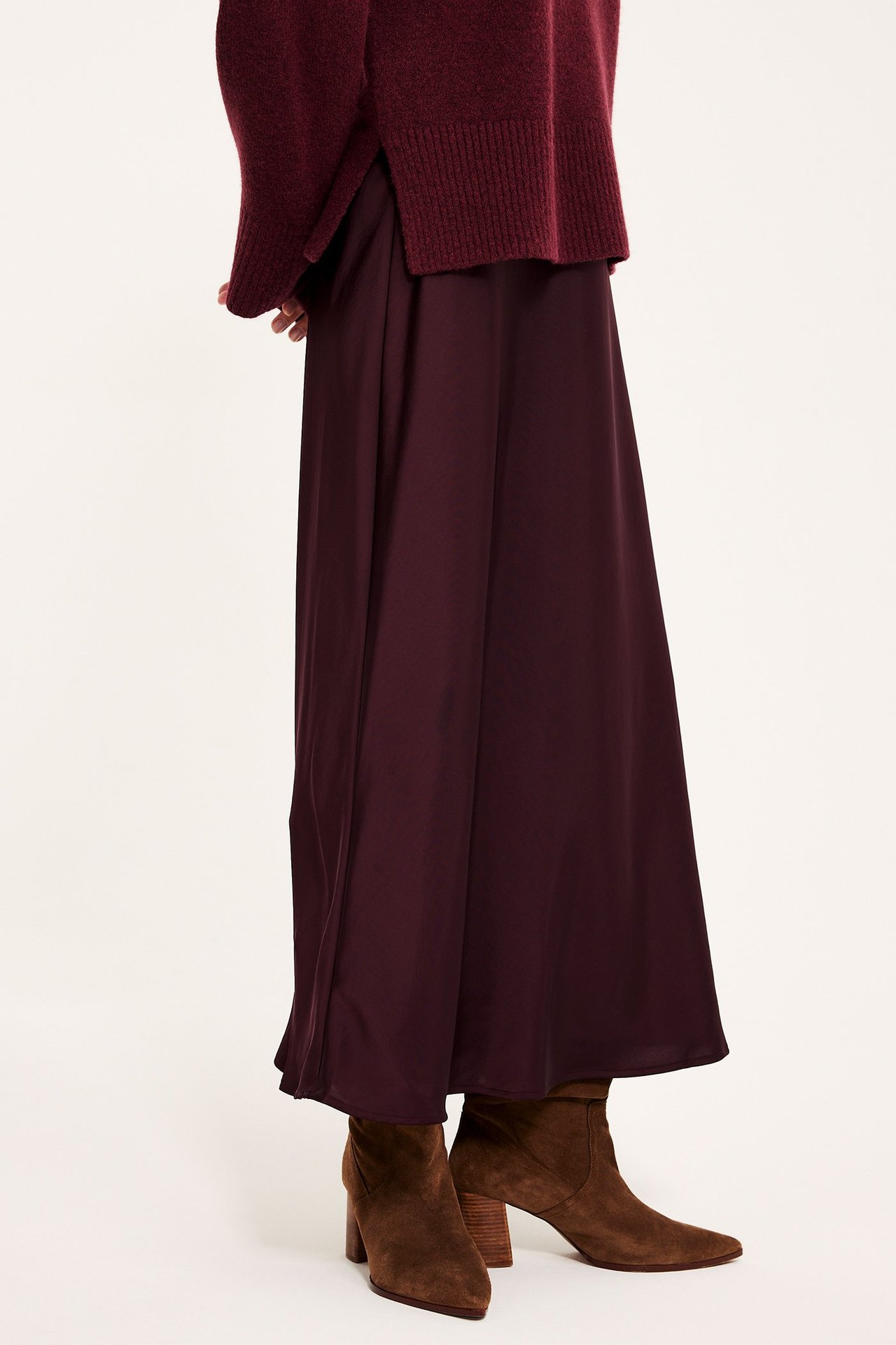 Sivukuva naisesta, jolla on viininpunainen levenevä satiinihame, yhteensopiva neulepusero ja ruskeat korkeakorkoiset nilkkurit.