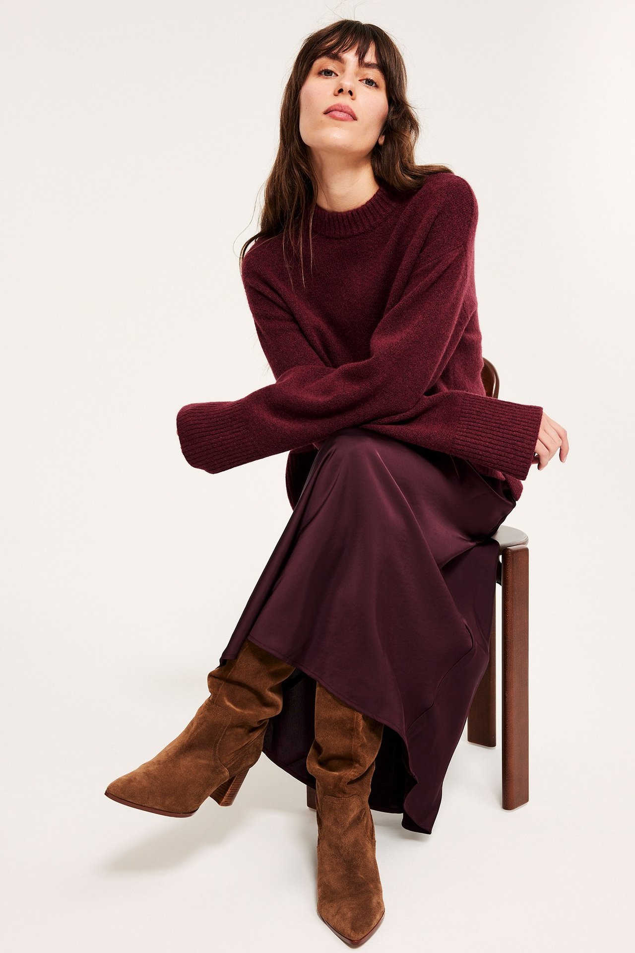 Edestä kuvattu nainen pitkässä, levenevässä viininpunaisessa satiinihameessa, neuleessa ja ruskeissa mokkanahkasaappaissa.