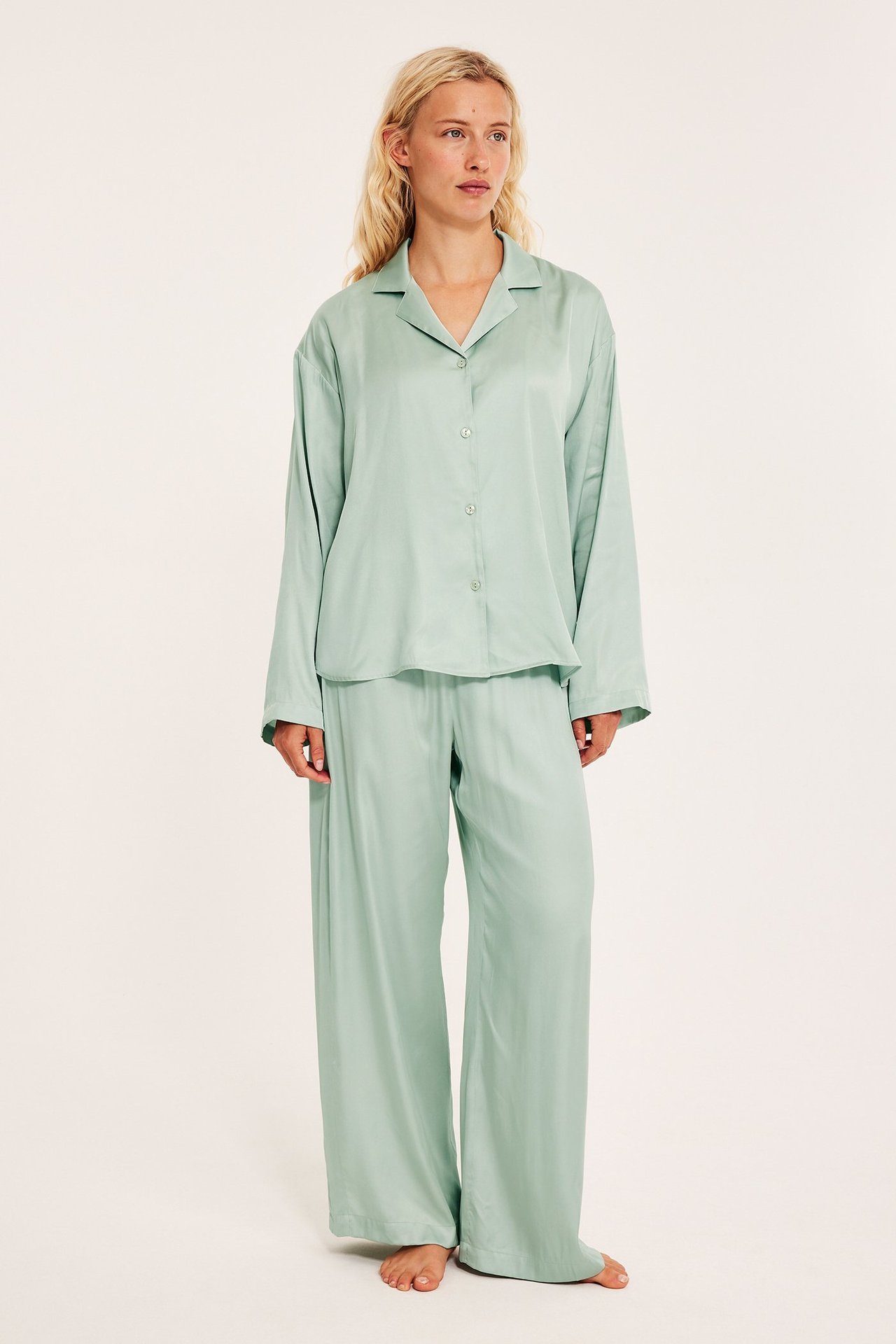 Edestä: Nainen vaaleanvihreässä satiinipyjamassa, löysä napitettava paita ja leveälahkeiset housut.