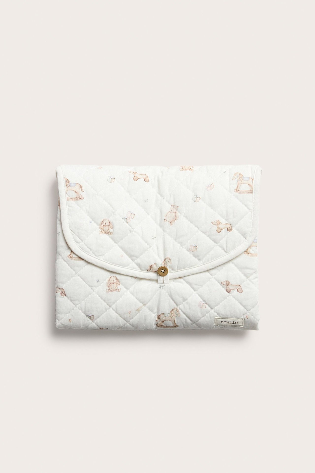 Frontvy av en offwhite quiltad vikbar skötbädd med leksaksmönster och knappstängning.