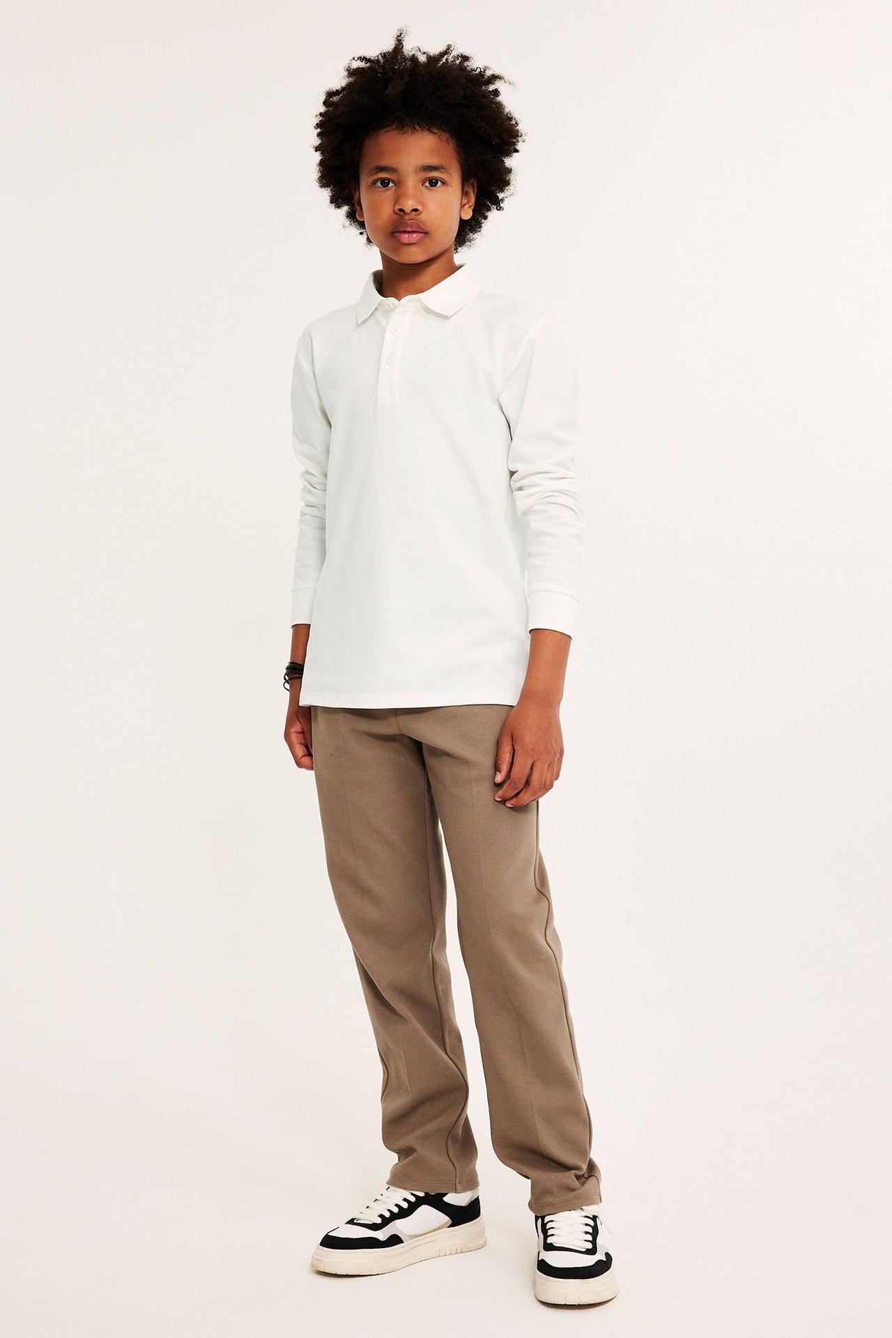 Edestä: Lapsi yllään luonnonvalkoinen pitkähihainen pikeepaita kolmella napilla, ruskeat housut ja lenkkarit.