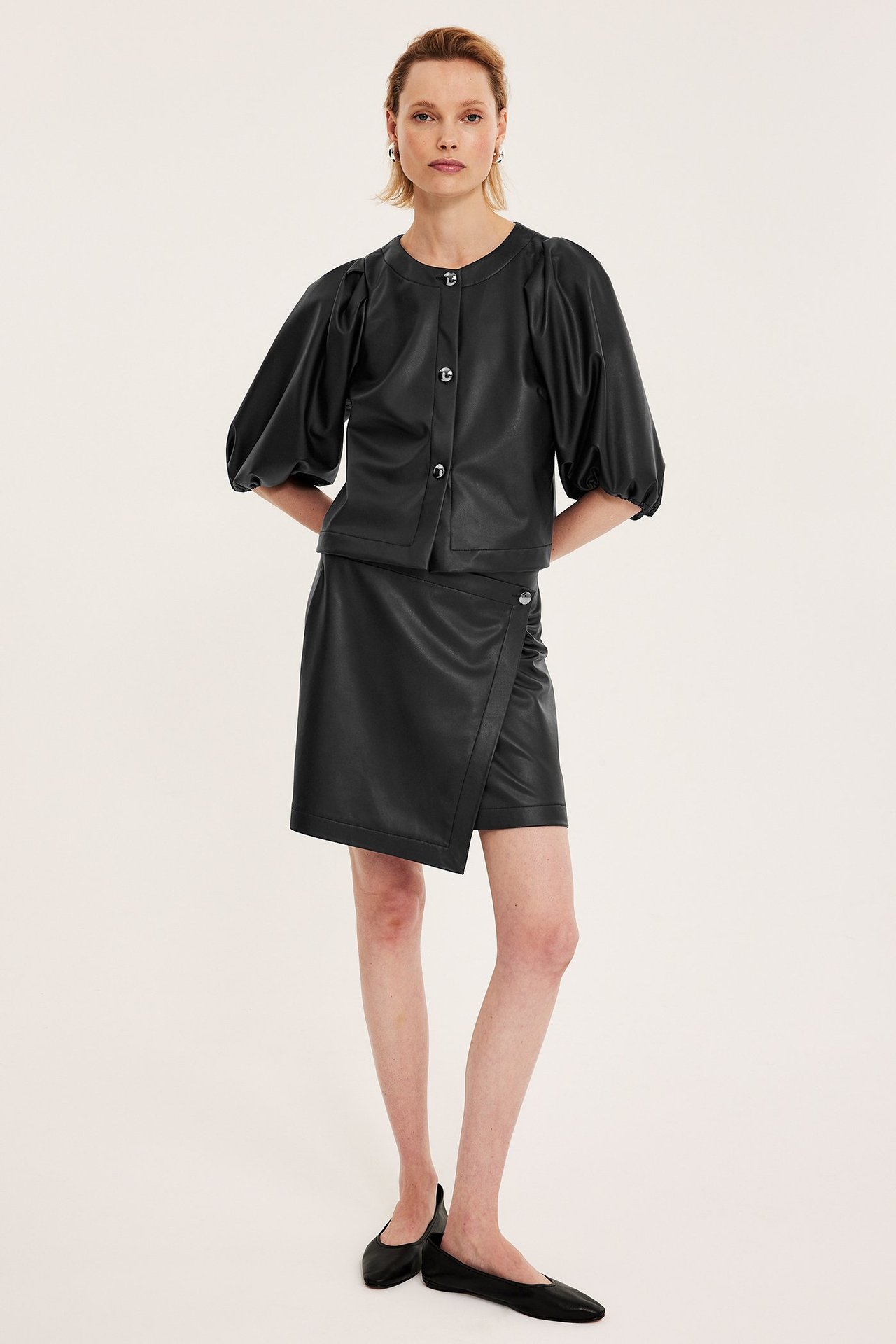 Framifrån: Kvinna i svart PU-blus med puffärmar, knappar, svart omlottkjol och svarta ballerinaskor.