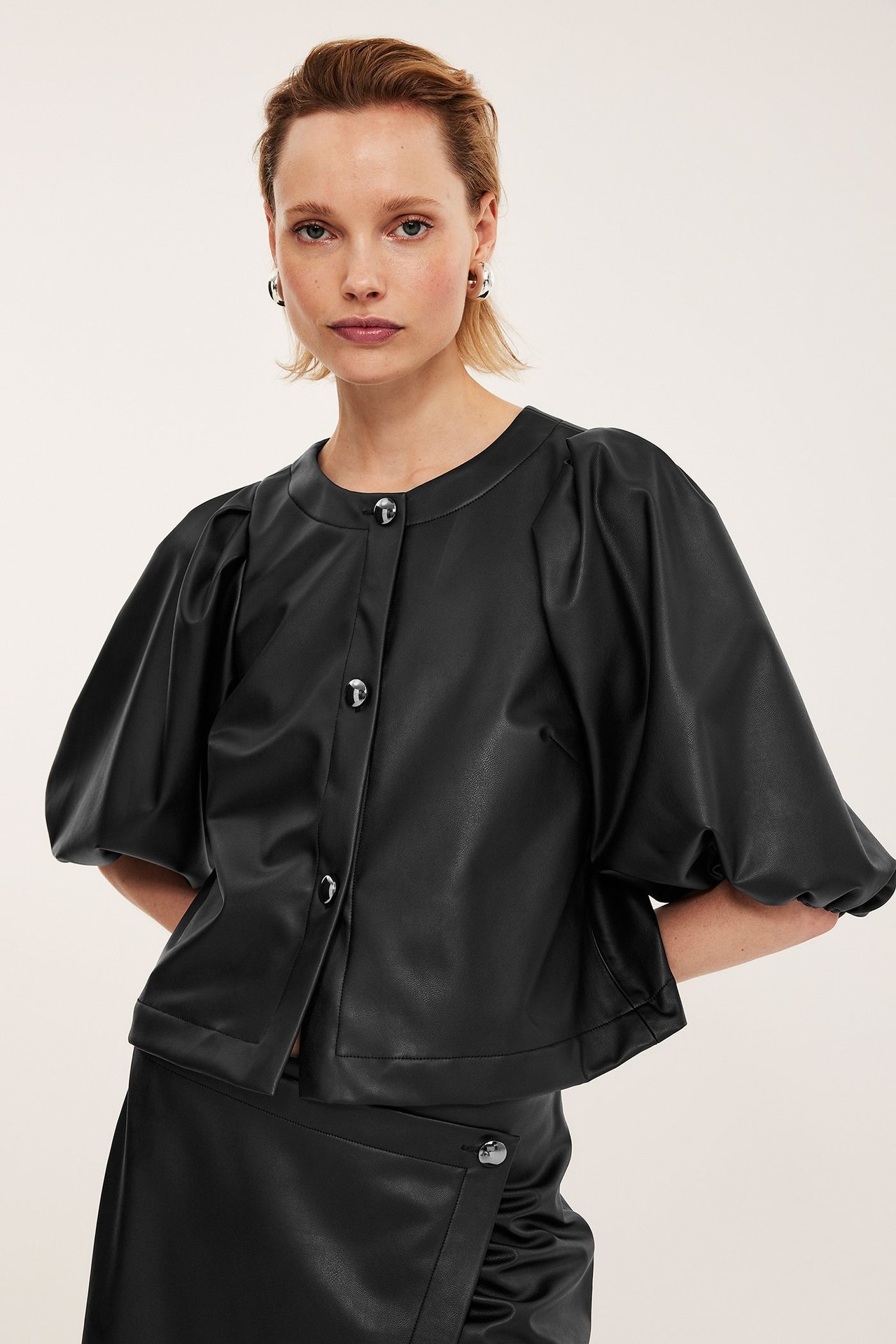 Widok z przodu: kobieta w czarnej bluzce PU z bufiastymi rękawami, srebrnymi guzikami i spódnicy PU.