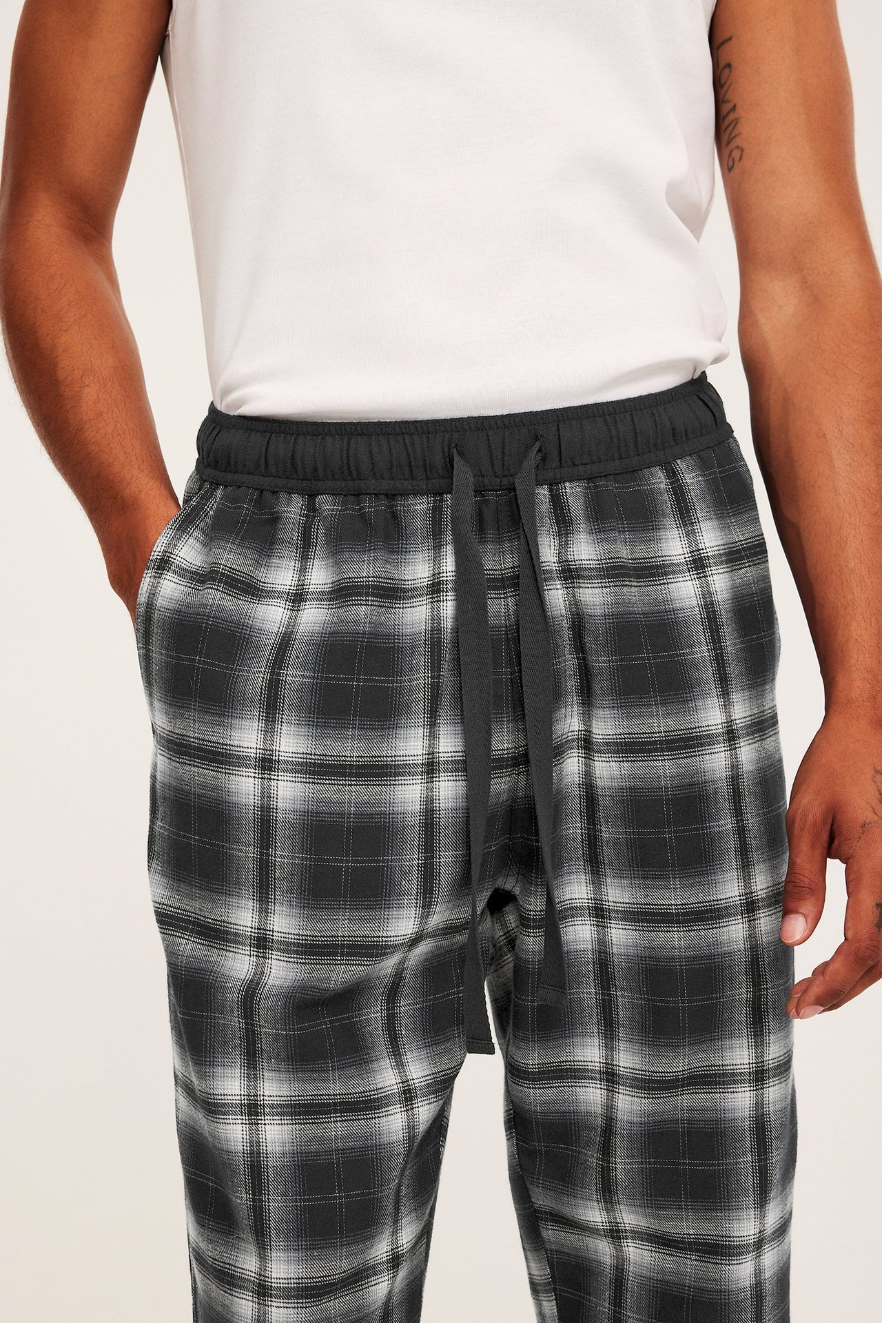 Lähikuva edestä: Mies mustavalkoisissa ruudullisissa flanellihousuissa, joissa kiristysnyöri, ja valkoinen t-paita.