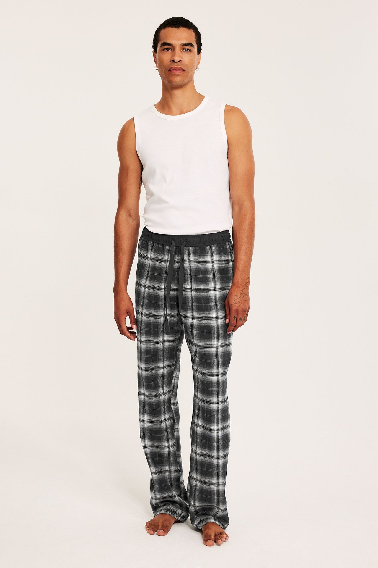 Edestä: mies musta-harmaa-valkoisissa ruudullisissa flanellipyjamahousuissa ja valkoisessa topissa.