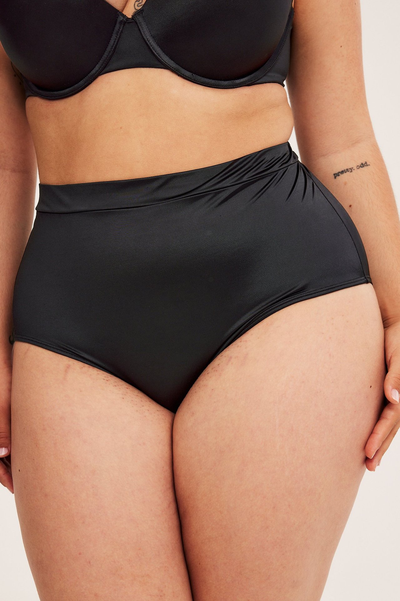 Närbild framifrån på en kvinna i svarta glansiga högmidjade brieftrosor och en svart BH.