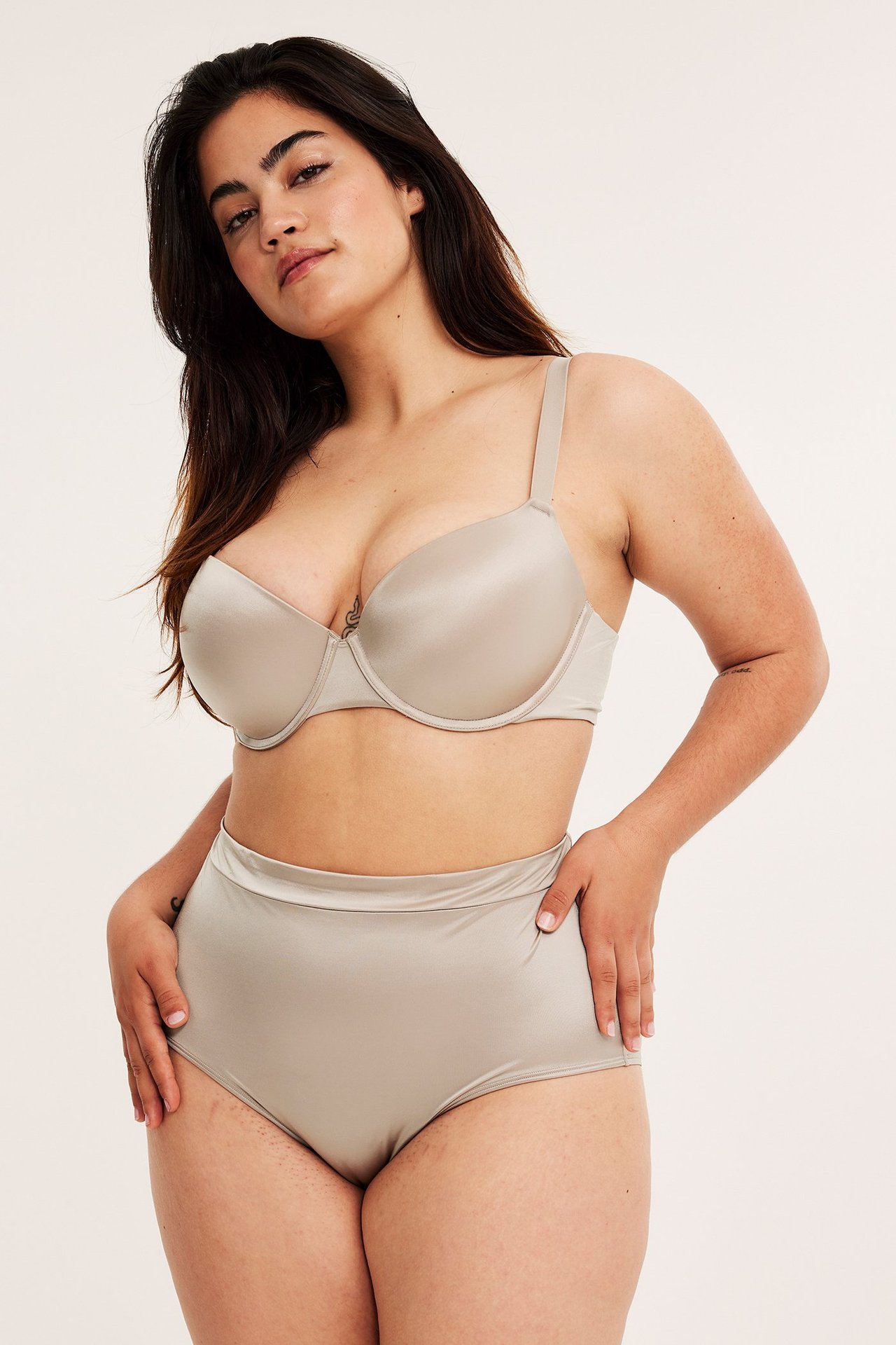 Frontbilde av en kvinne i en lys beige glinsende høy truse og en matchende BH.