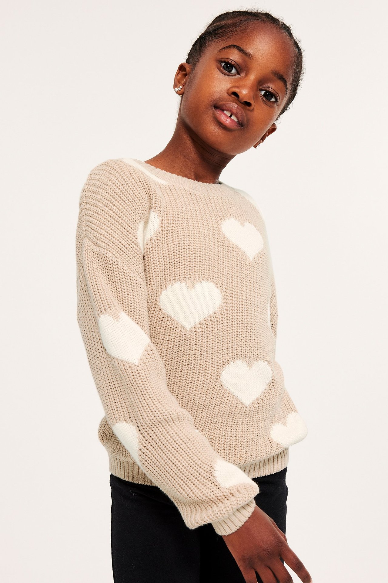 Widok z przodu dziecka w beżowym grubym swetrze z białymi sercami żakardowymi i czarnych spodniach.