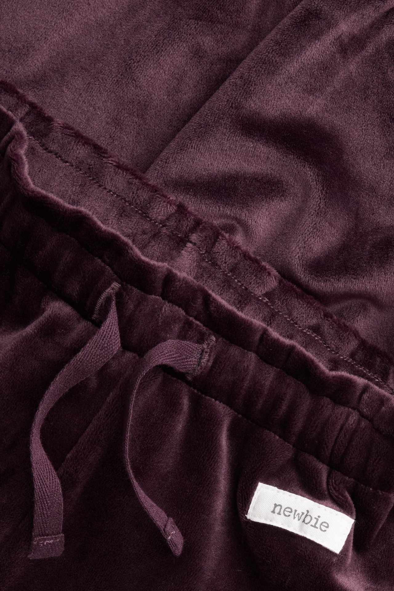 Zbliżenie liliowych welurowych spodni dresowych z elastycznym pasem, sznurkiem i białą metką "newbie".