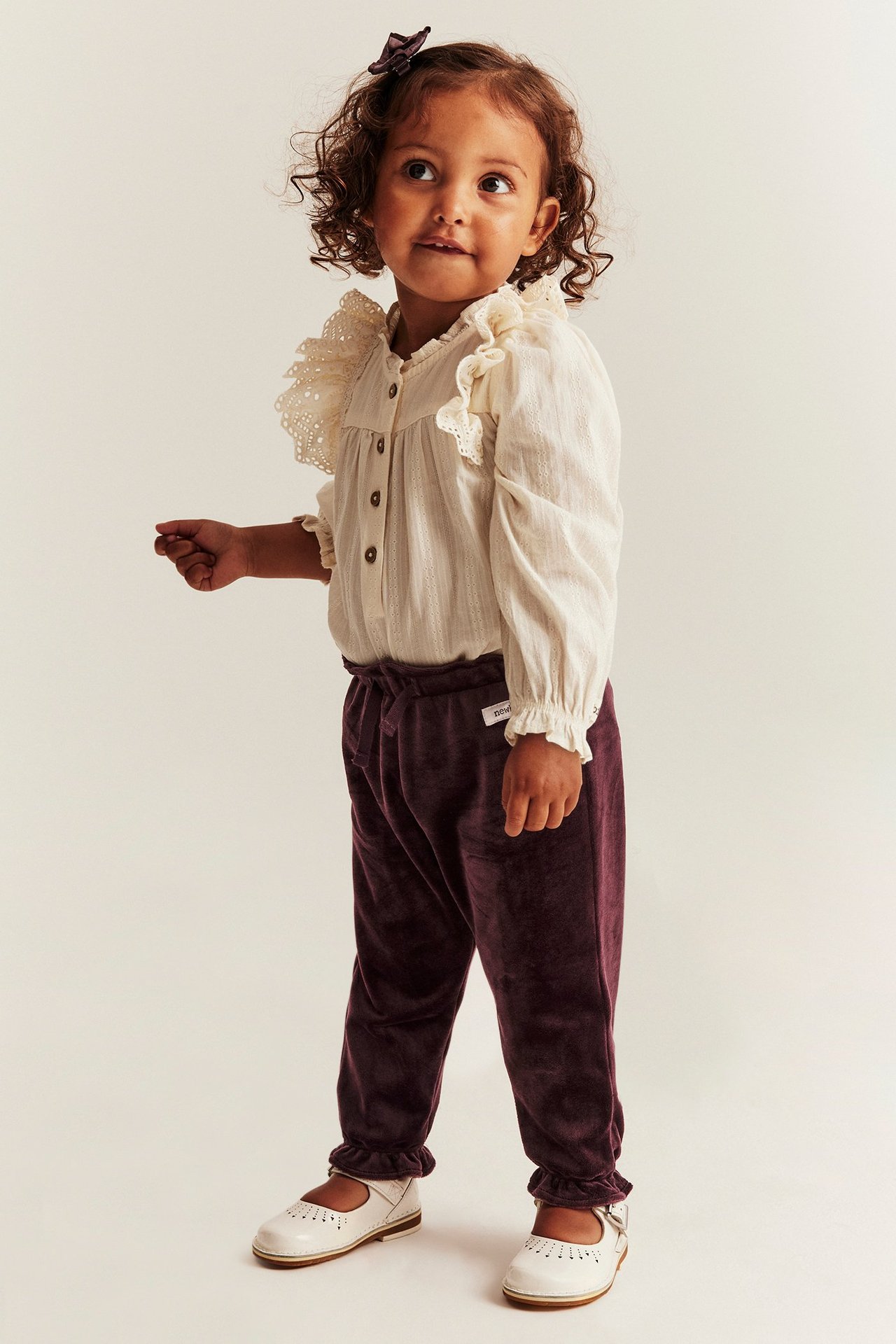 Widok z przodu: Dziecko w ciemnofioletowych welurowych spodniach dresowych, kremowej bluzce, białych butach, ciemnej spince.