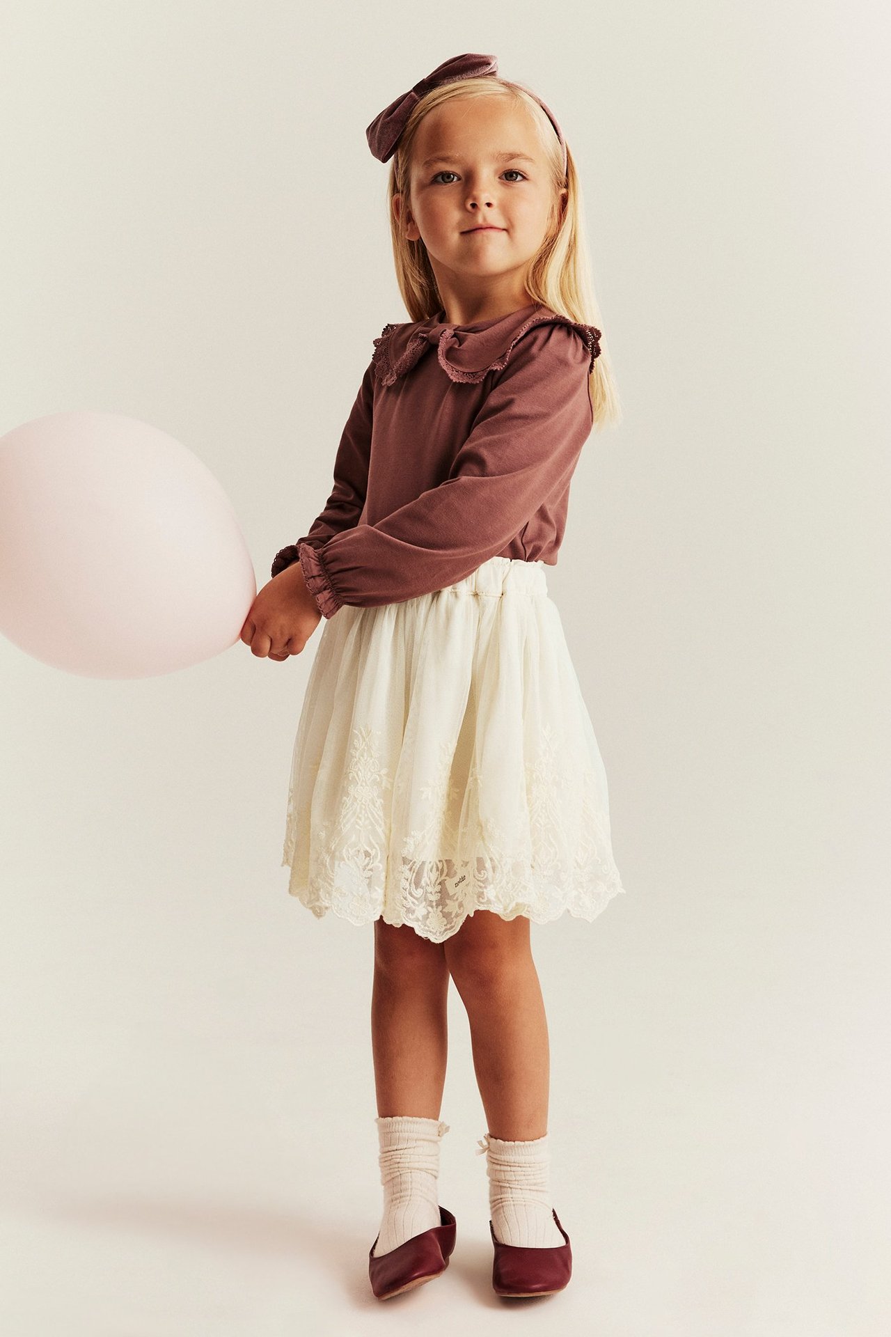 Barn i ljusbeige broderad meshkjol, brun tröja, rosett, gräddvita strumpor, röda ballerinaskor, håller ballong.