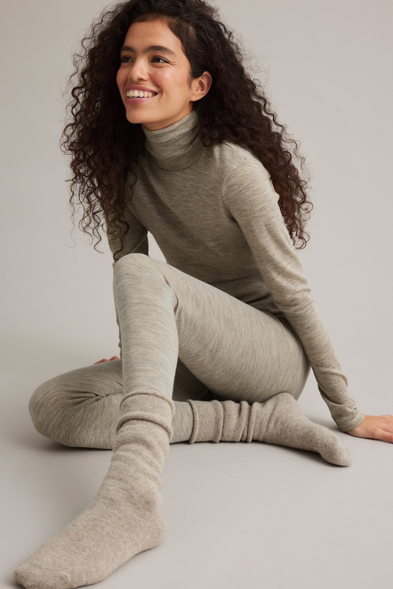 Side-frontbilde av smilende kvinne i beige melert merinoull-genser, matchende leggings og sokker.
