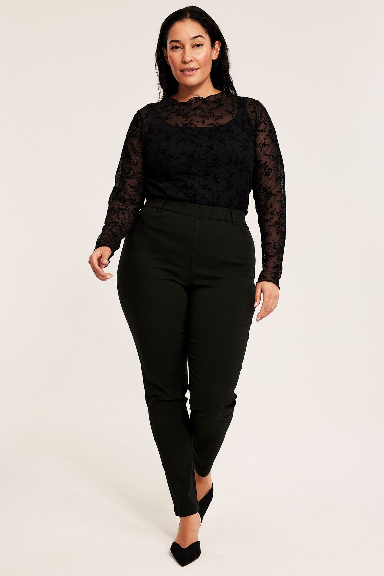 Kobieta w czarnych treggingsach z pętlami na pasek, w czarnej bluzce i szpilkach.