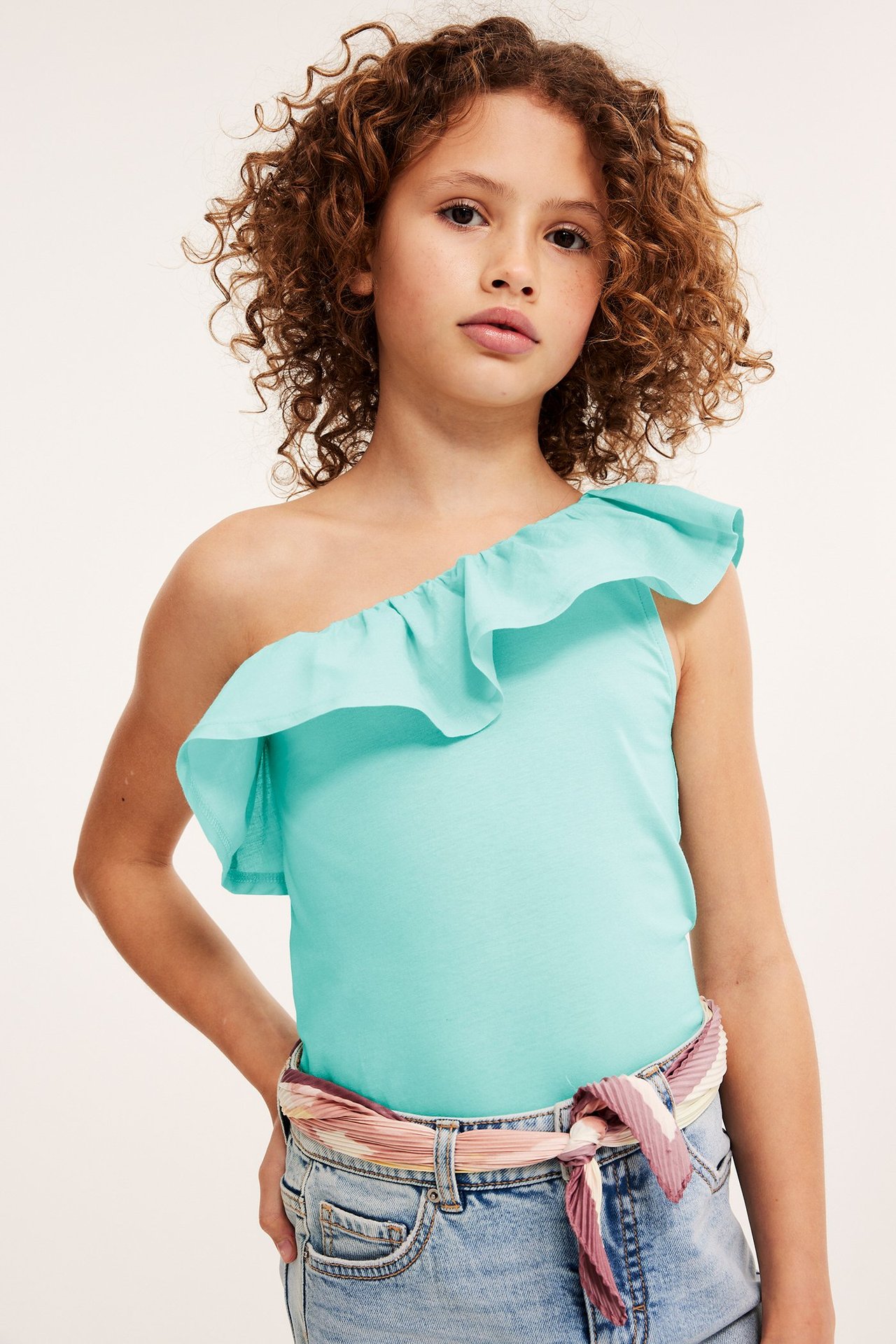 Barn bär en ljus turkos one-shoulder topp med volang, ljusblå denimshorts och ett plisserat sjalbälte.