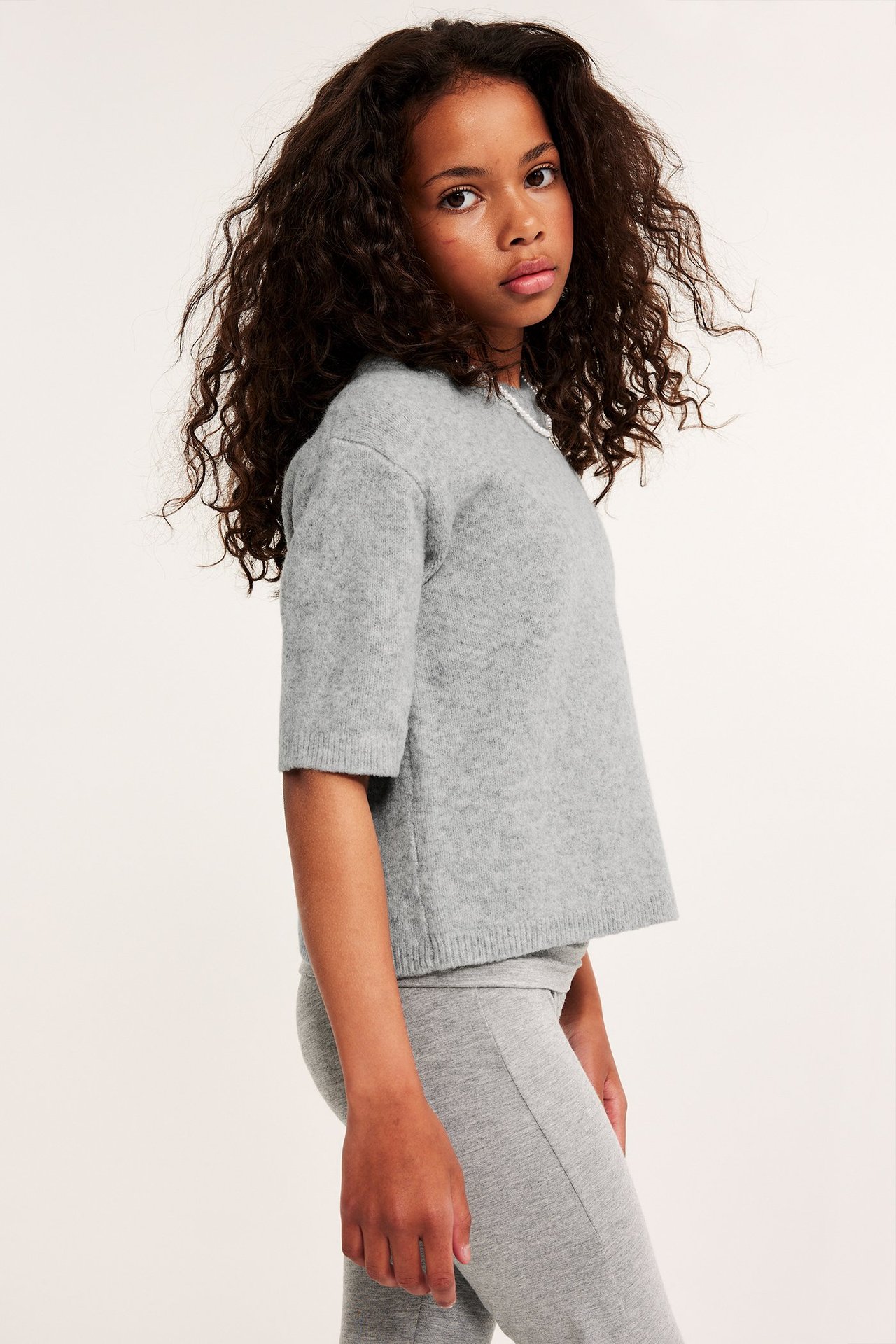 Sidovy av ett barn i en ljusgrå melerad finstickad kortärmad t-shirt och ljusgrå leggings.