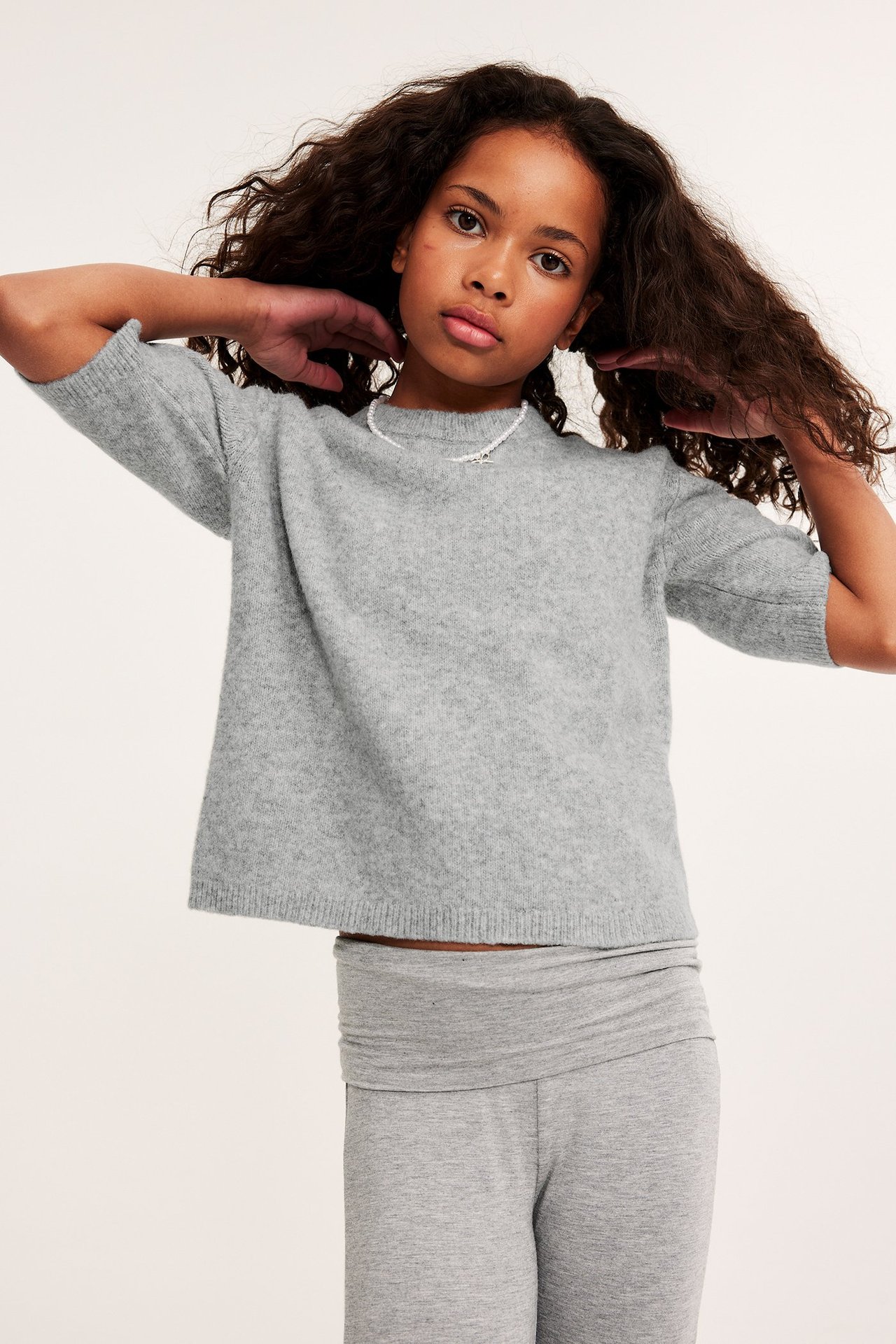 Frontvy: Barn i ljusgrå melerad kortärmad stickad tröja, ljusgrå leggings och pärlhalsband.