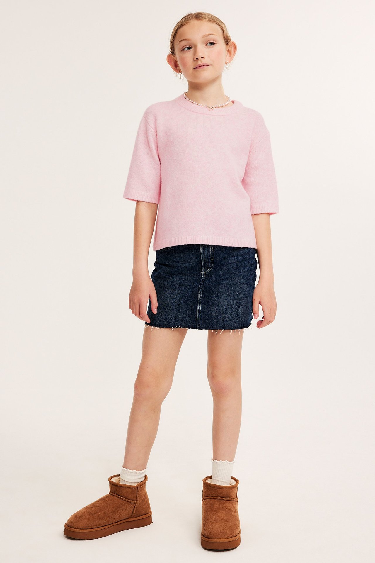 Framsida: Flickbarn i rosa melerad kortärmad stickad t-shirt, mörkblå jeanskjol och bruna stövlar.