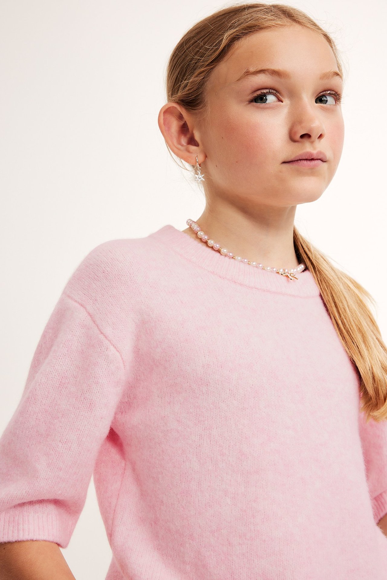 Närbild på ett barn i en ljusrosa melerad kortärmad stickad tröja, ett pärlhalsband och stjärnörhängen.
