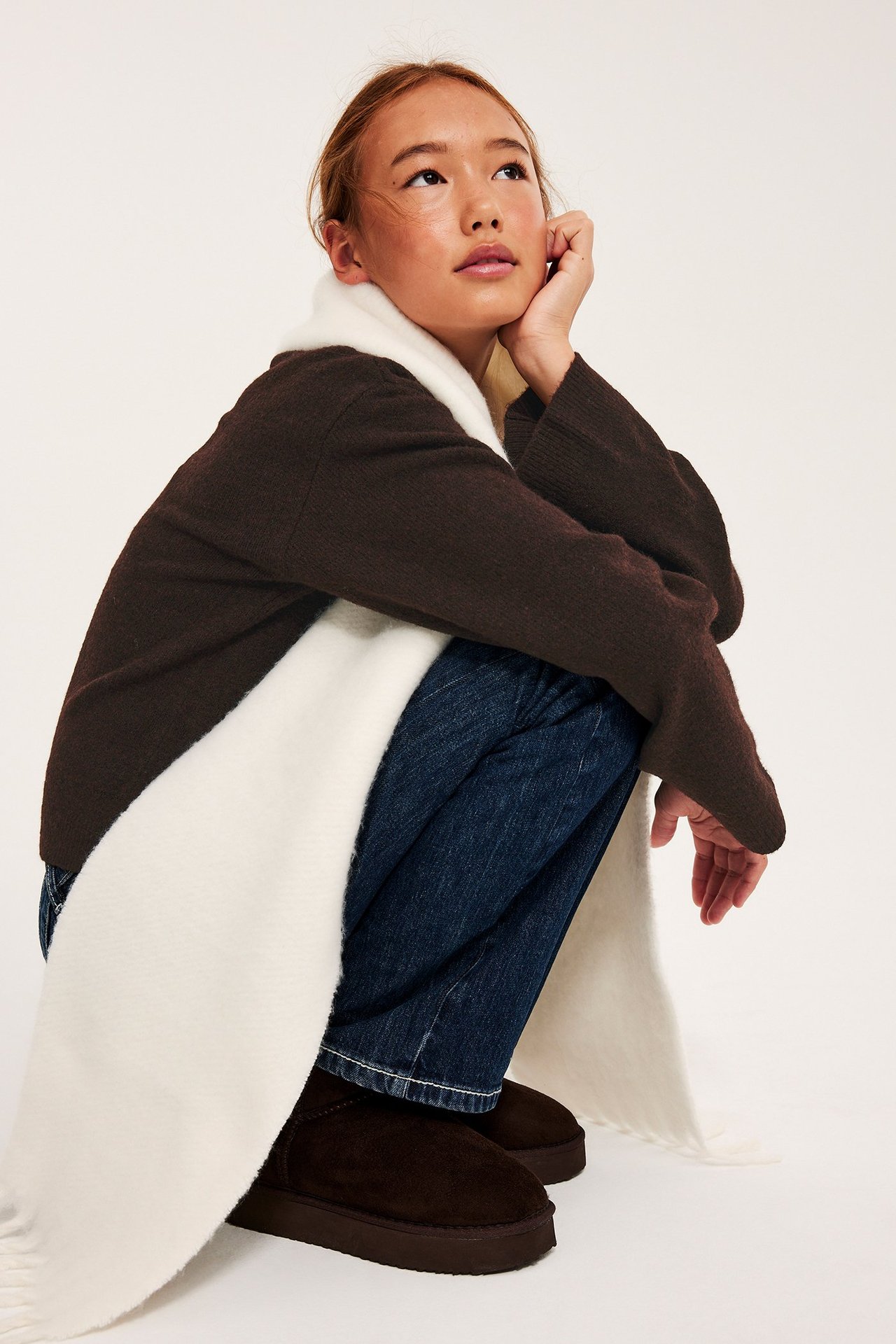 Sned framifrån vy av en kvinna i brun finstickad tröja, vit halsduk, blå jeans och bruna stövlar.