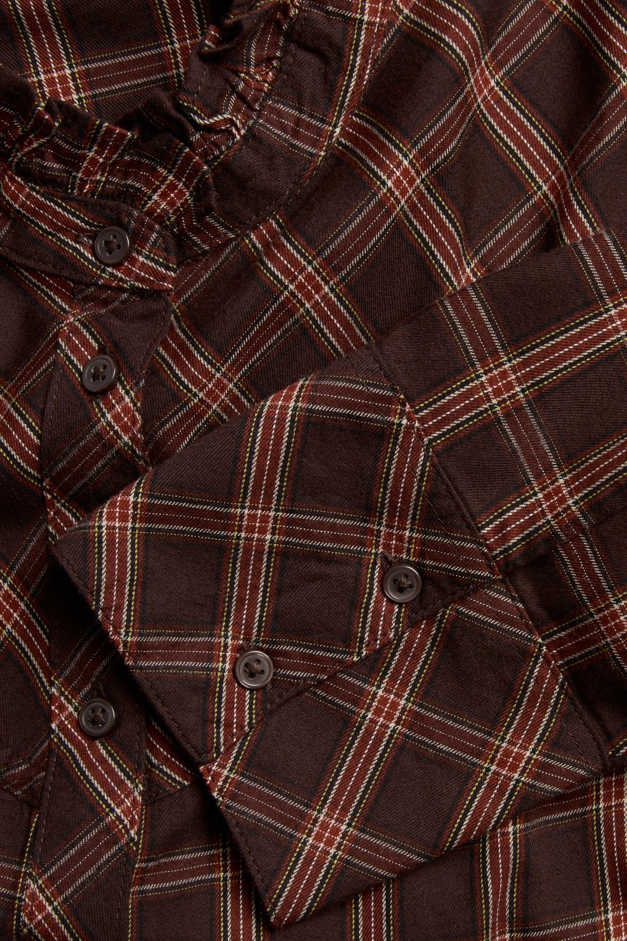 Zbliżenie na bordową bluzkę w kratę z falbankowym dekoltem i zapinanymi mankietami.