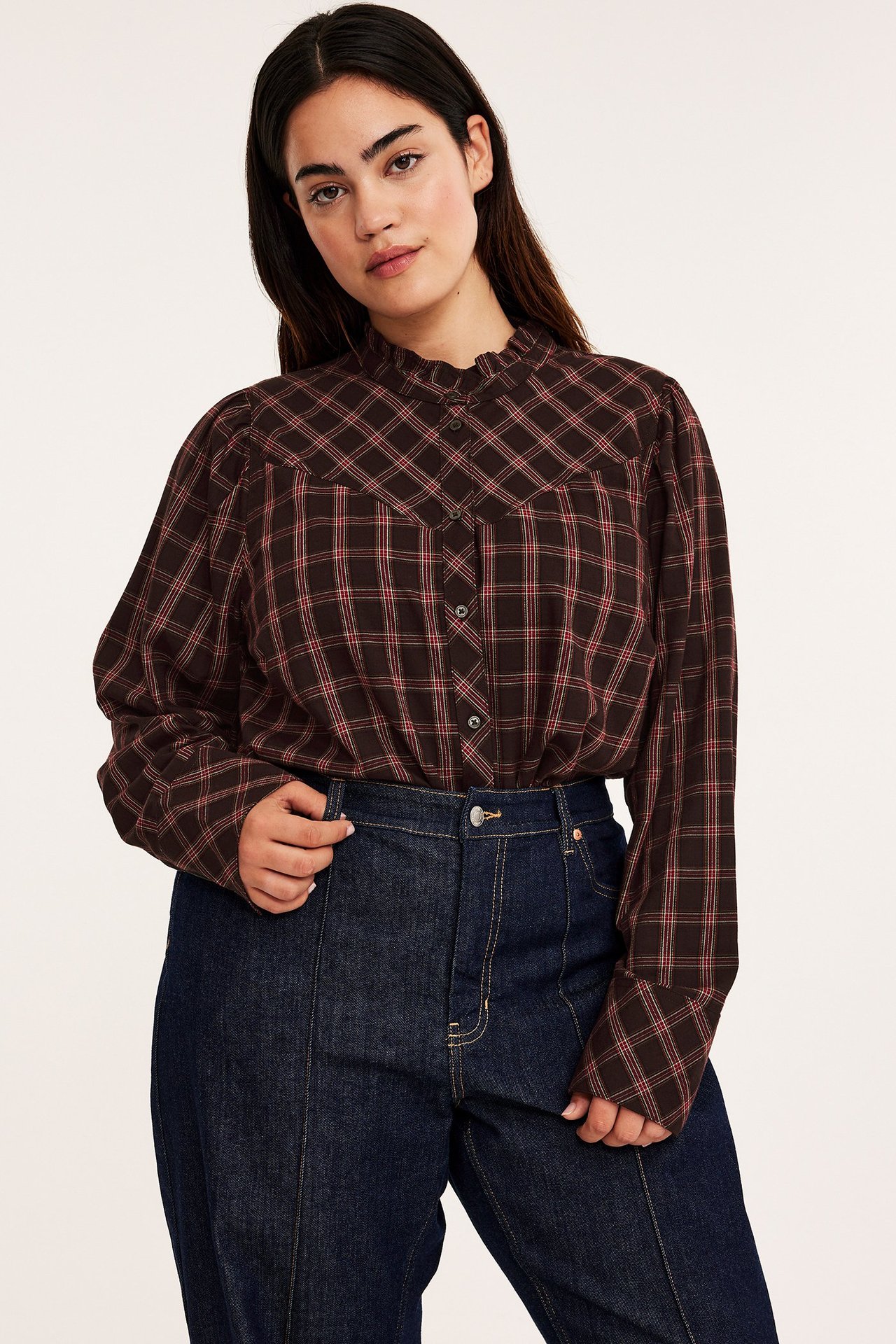 Widok z przodu: kobieta w ciemnobrązowej bluzce w kratę z falbanką i ciemnoniebieskich dżinsach.