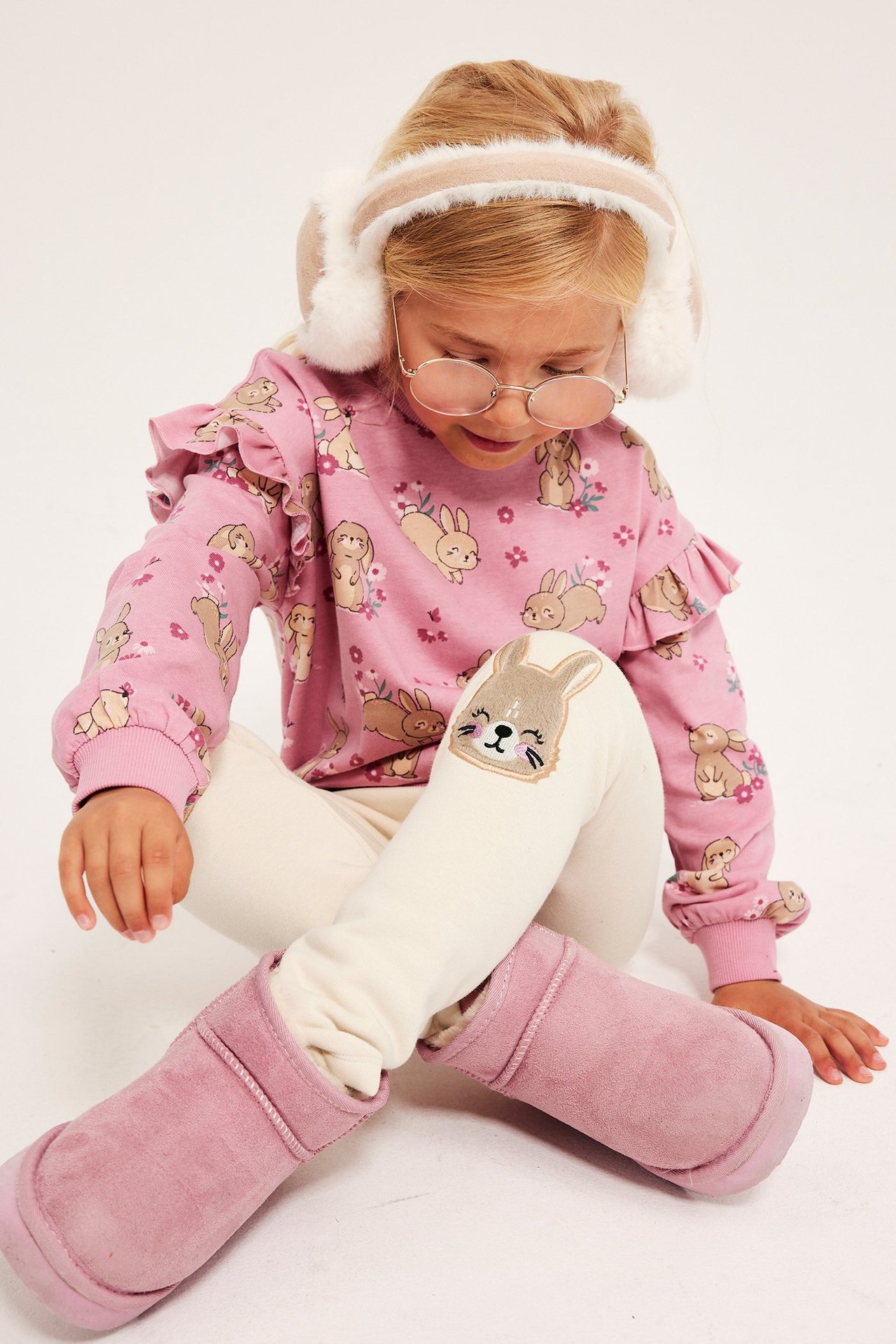 Forfra: Et barn i lysebeige kanin-applikasjonsleggings, rosa kaninmønstret topp, beige øreklokker og rosa støvler.
