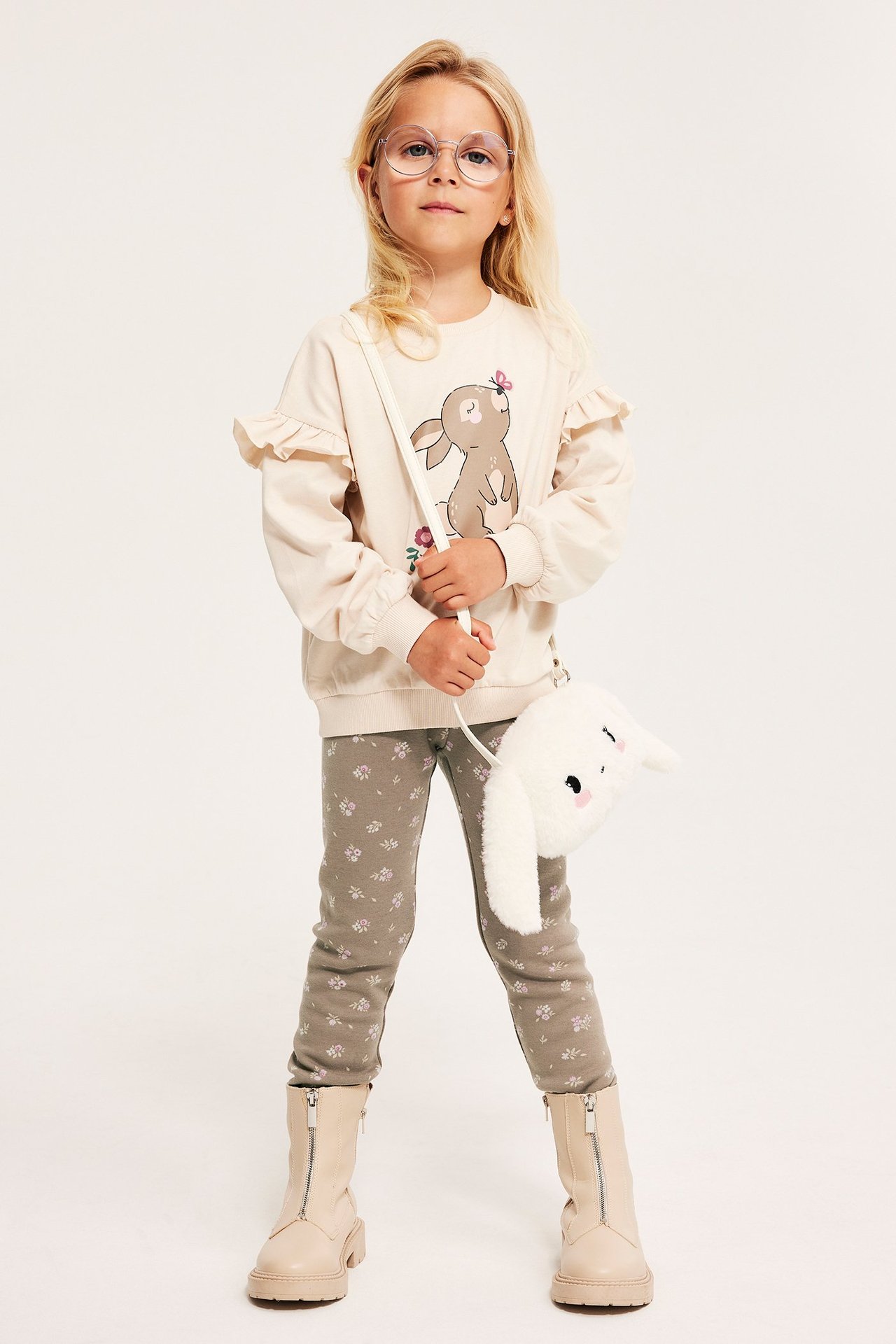 Widok z przodu: Dziecko w beżowych legginsach w kwiaty, beżowej bluzie, beżowych butach, z pluszową torbą-zającem.