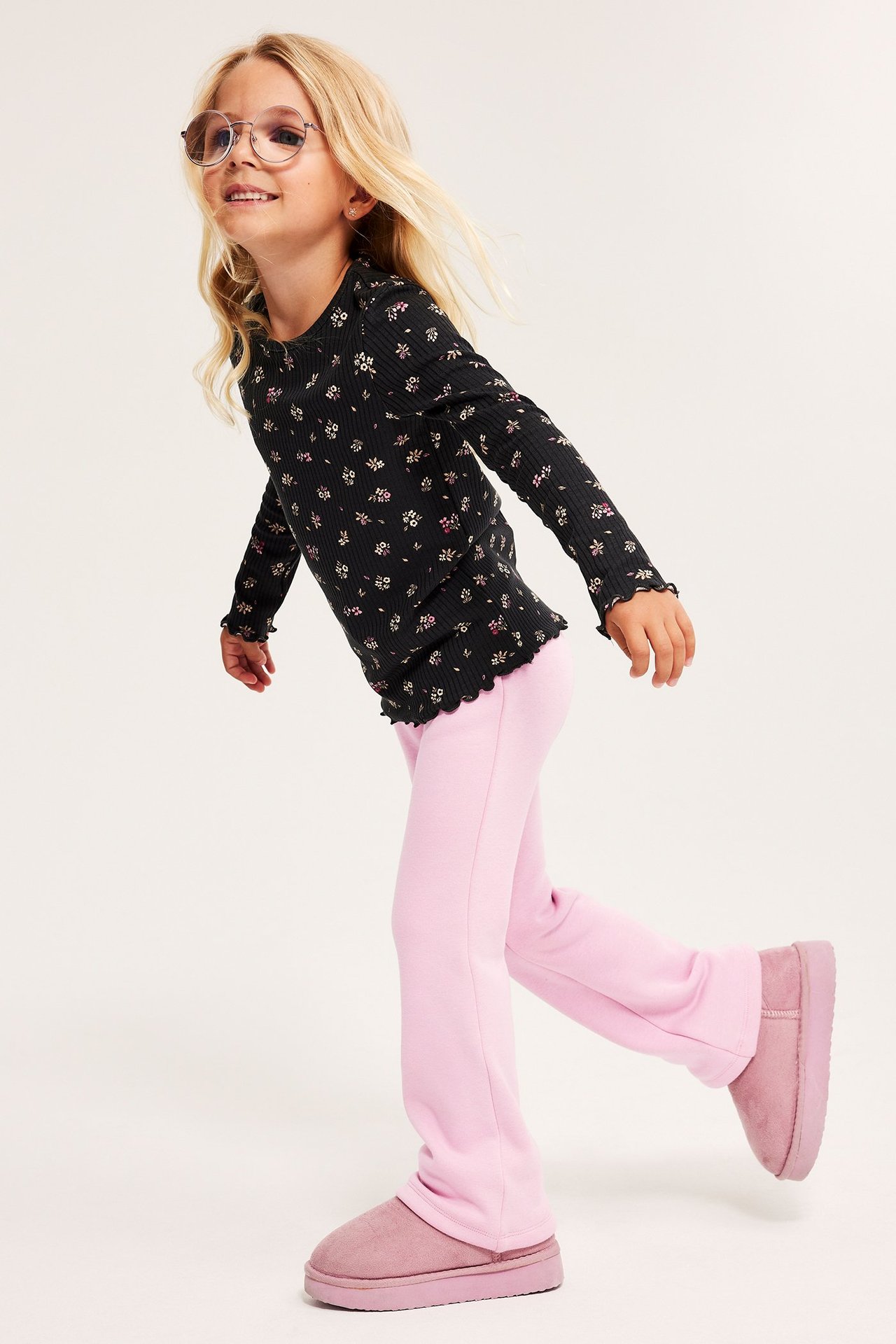 Widok z boku dziecka w jasnoróżowych legginsach dzwonach, czarnej bluzce w kwiaty i różowych butach.